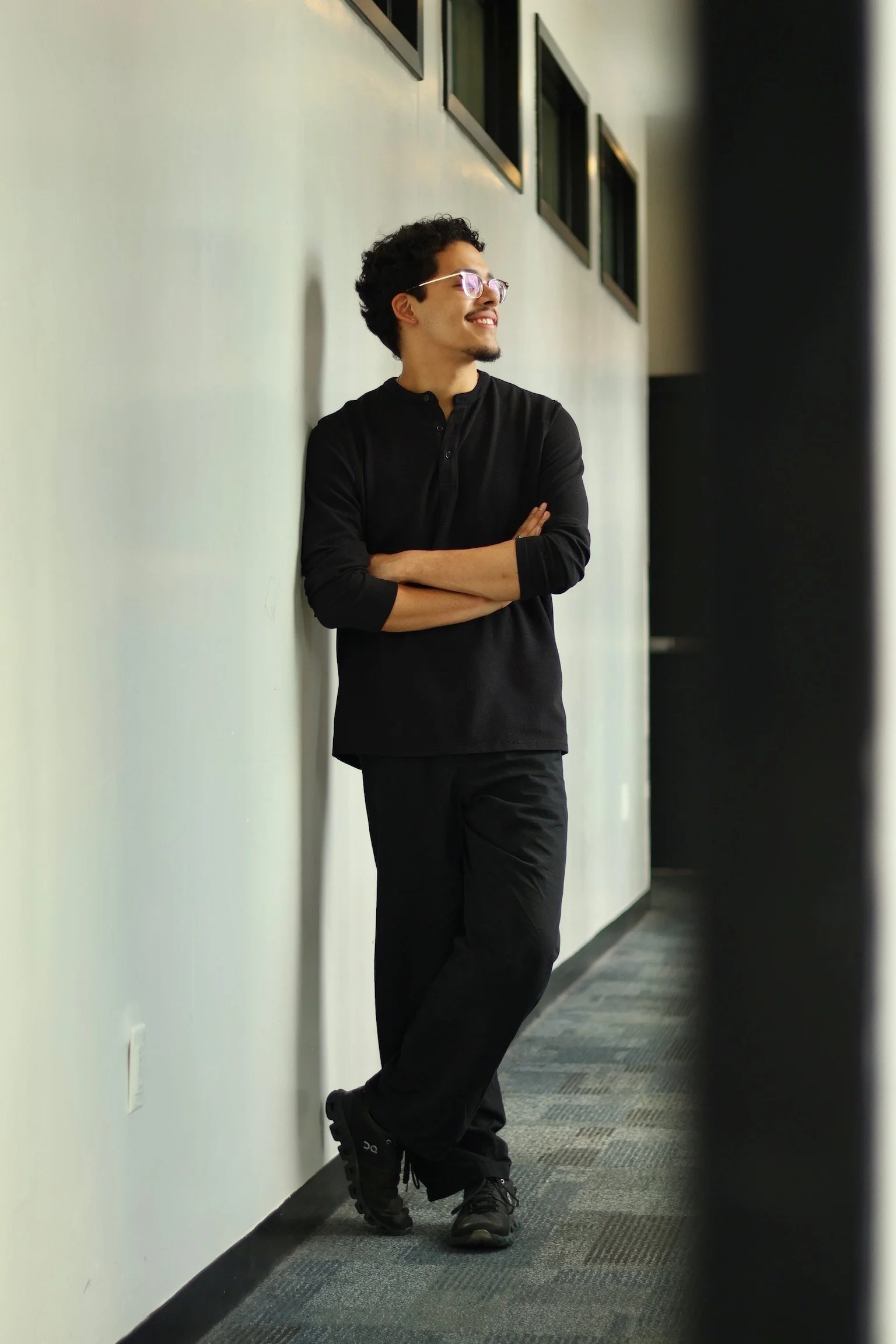 A young man with dark curly hair, wearing glasses, a black shirt with rolled-up sleeves, black pants, and black sneakers, stands with arms crossed, leaning against a white wall with his right shoulder, smiling and looking to the left.