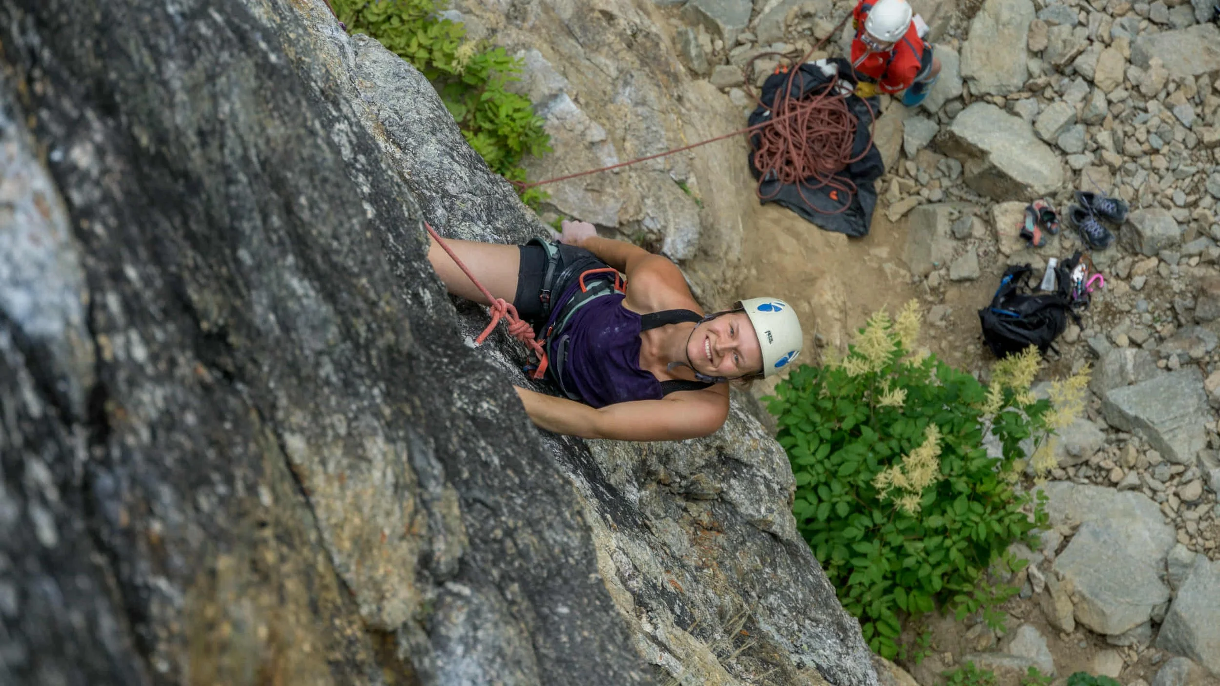 En kvinde klatrer op ad en klippevæg med hjelm og reb, smilende og ser op mod kameraet, omgivet af klippeblokke og grønne planter.