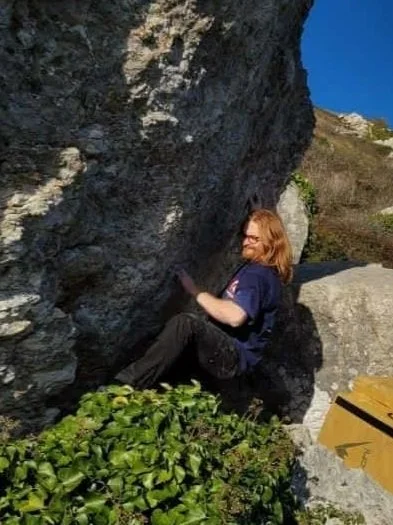 En person med brunt hår sidder under en stor klippe på en fjeldsti, omgivet af grønne planter og klipper, solrigt vejr.