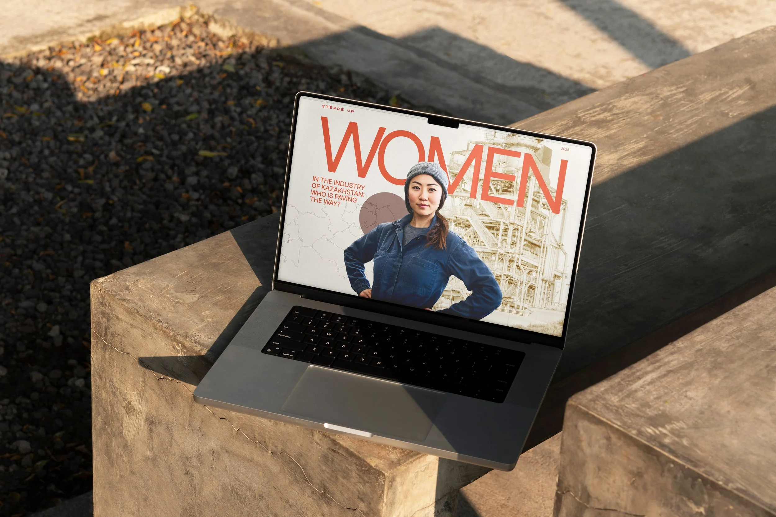 A laptop resting on a stone ledge outdoors displaying a magazine cover with a woman wearing a gray beanie and blue jacket, against an industrial background with text about women in the Kazakhstan industry.