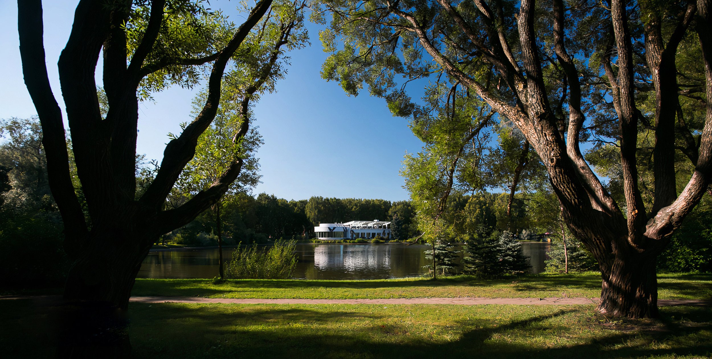 A peaceful park scene featuring large trees with green leaves, a calm lake reflecting the sky and surrounding trees, and a modern white building in the distance under a clear blue sky.