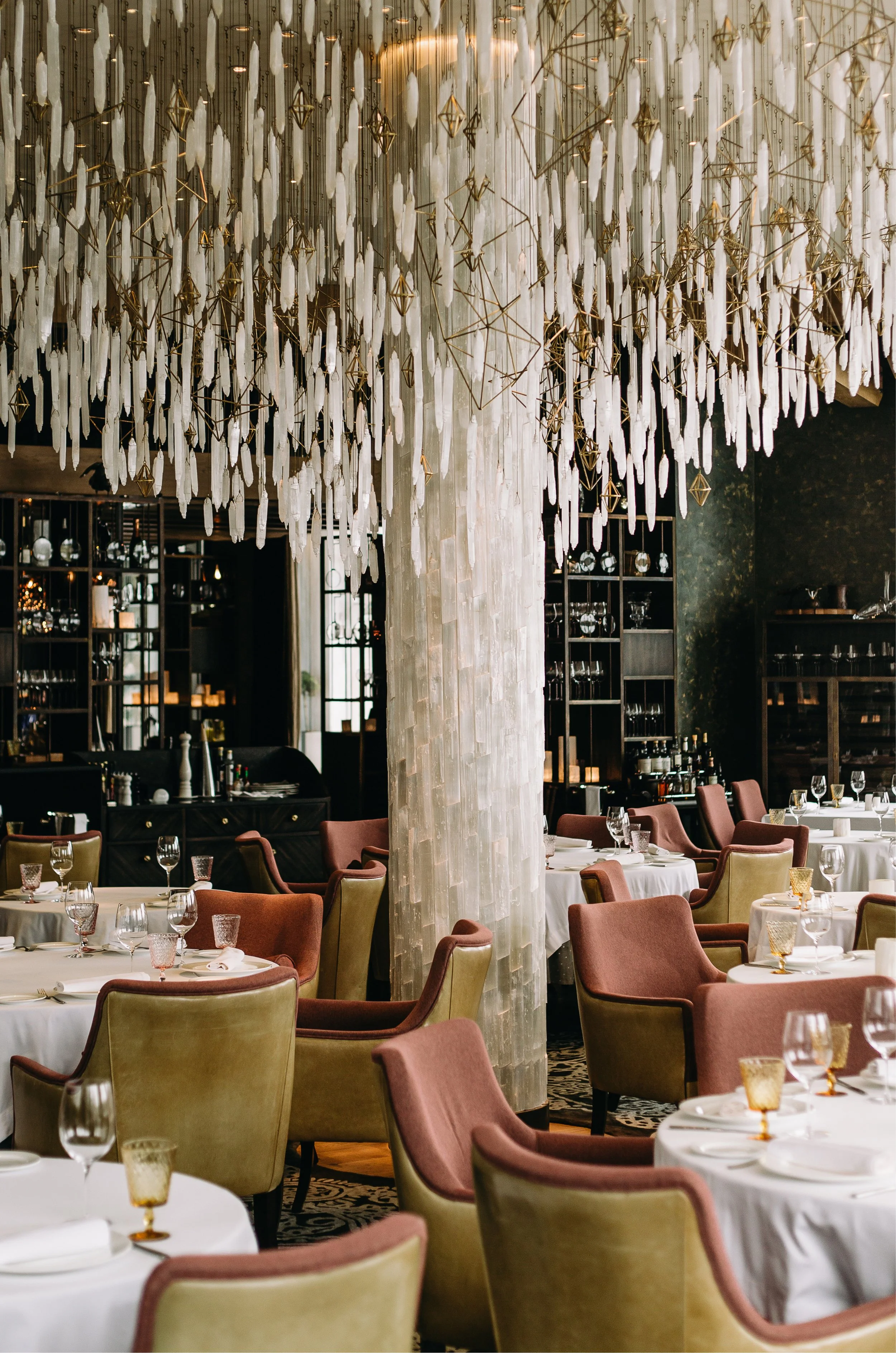 Elegant restaurant interior with white tablecloths, glassware, and upholstered chairs, featuring a decorative hanging installation with white feathers and gold geometric accents.