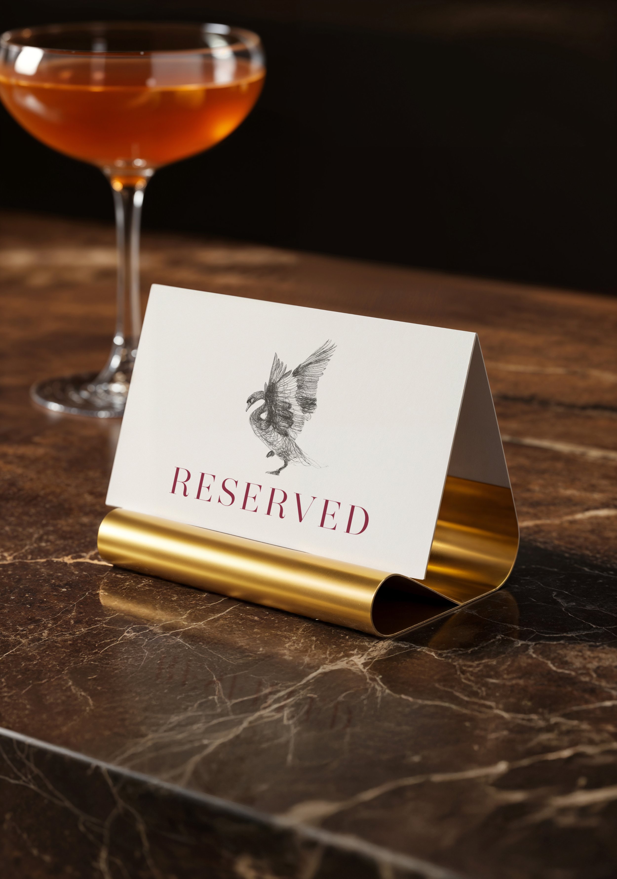 A reserved table sign with a sketch of a bird, placed on a dark marble table, with a glass of amber-colored drink in the background.