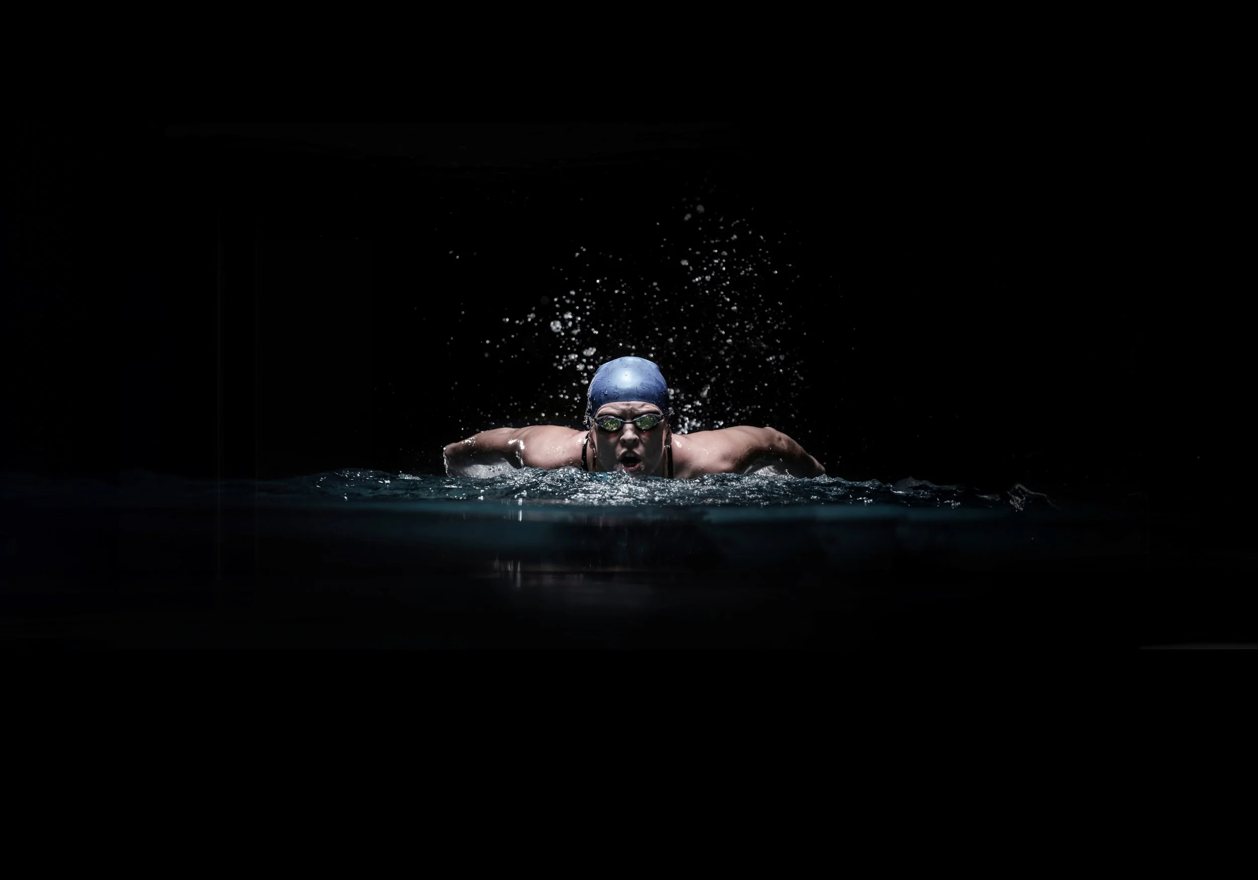 Swimmer with goggles and swim cap performing a butterfly stroke in a dark pool, with water splashing around.