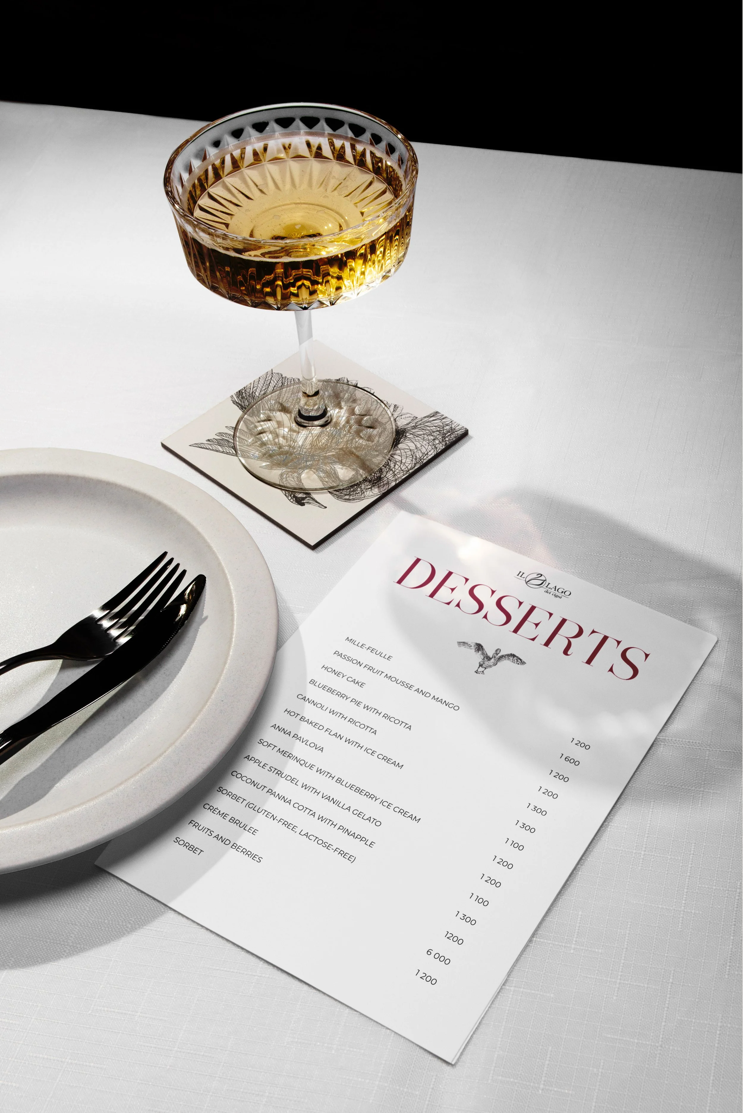 A glass of champagne on a white table with a dessert menu, silver fork and knife on a white plate. The menu is partially visible with desserts listed and prices.
