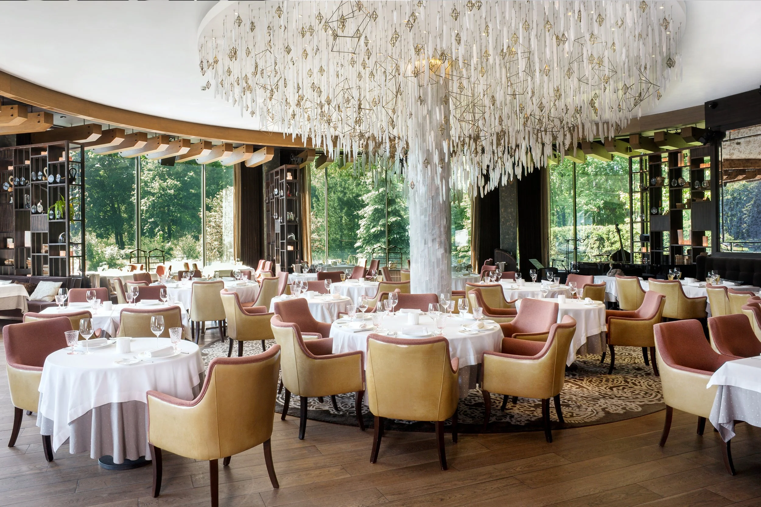 Elegant restaurant dining area with round tables covered in white tablecloths, surrounded by upholstered chairs in beige and pink tones. Large windows offer views of greenery outside. A grand, modern chandelier with gold accents hangs from the ceiling.