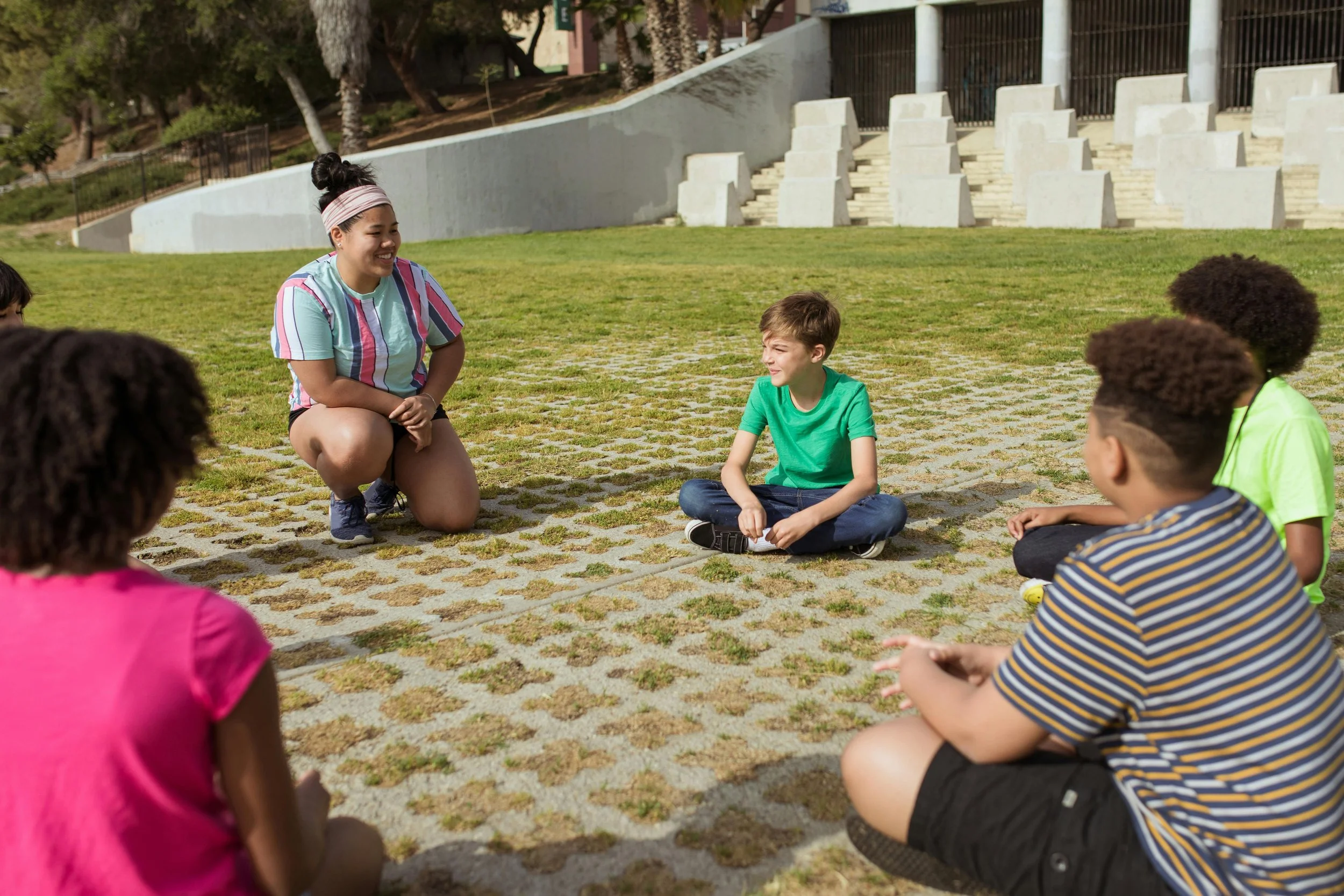 A group of children sitting on the grass in a semi-circle outdoors, engaging in a discussion or game, with a young woman crouching and smiling at them.