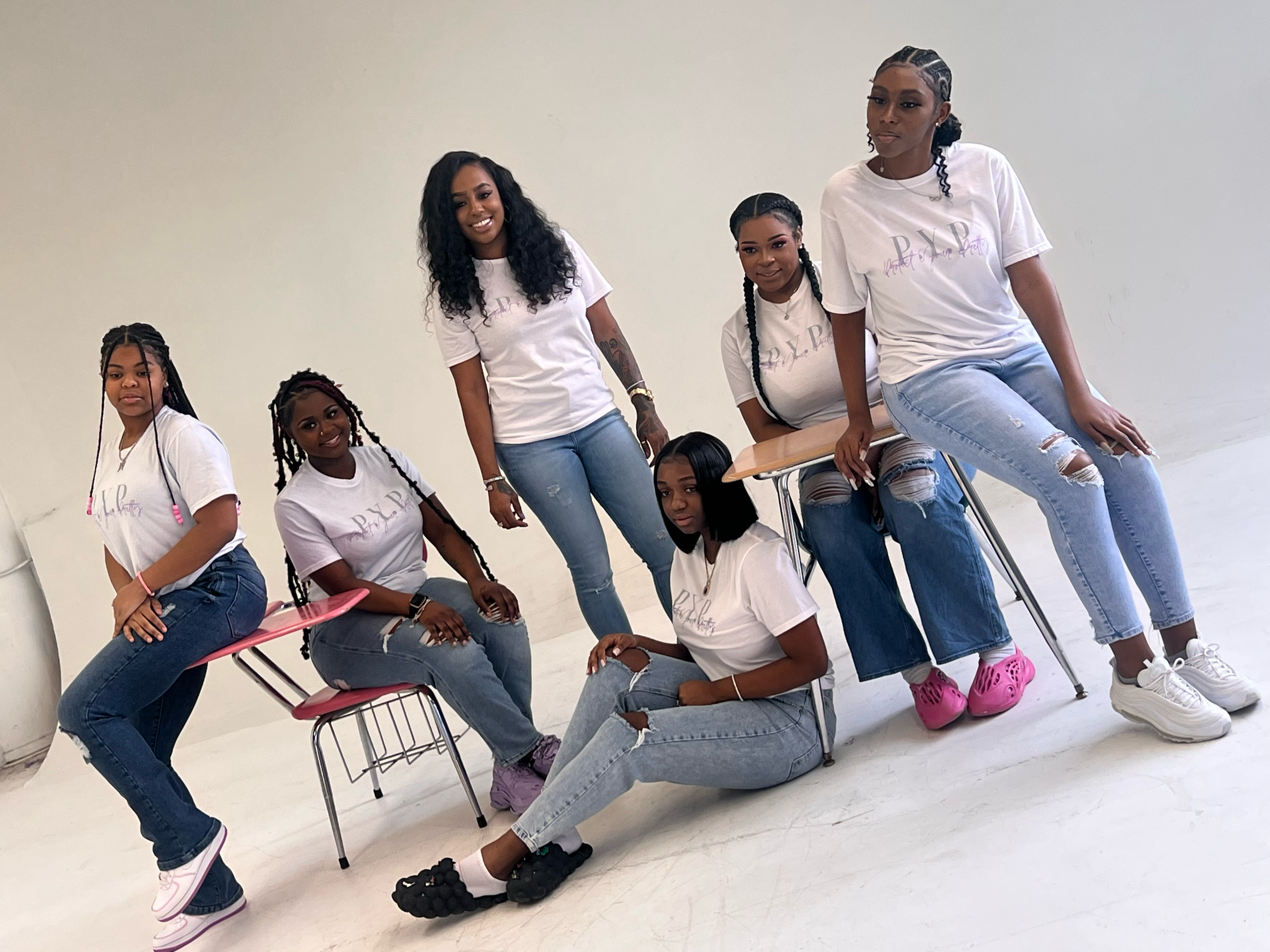 Seven women in white t-shirts with matching logos, sitting or standing on chairs and the floor in a white room.