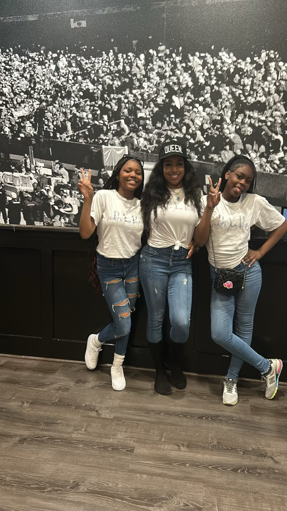 Three women standing together in front of a large black and white photo of a crowded event, all smiling and making peace signs with their hands. They are dressed casually, wearing jeans and white tops, with one wearing a "QUEEN" cap.