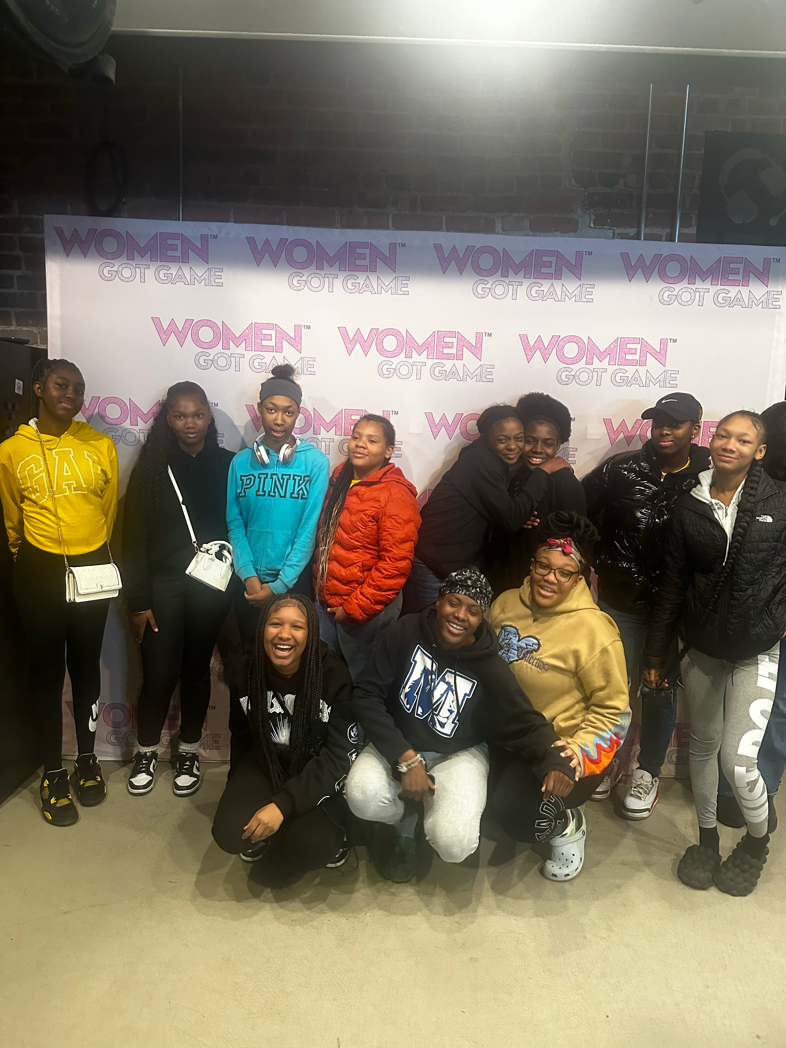 Group of women and girls posing in front of a backdrop with the text 'Women Got Game' multiple times.