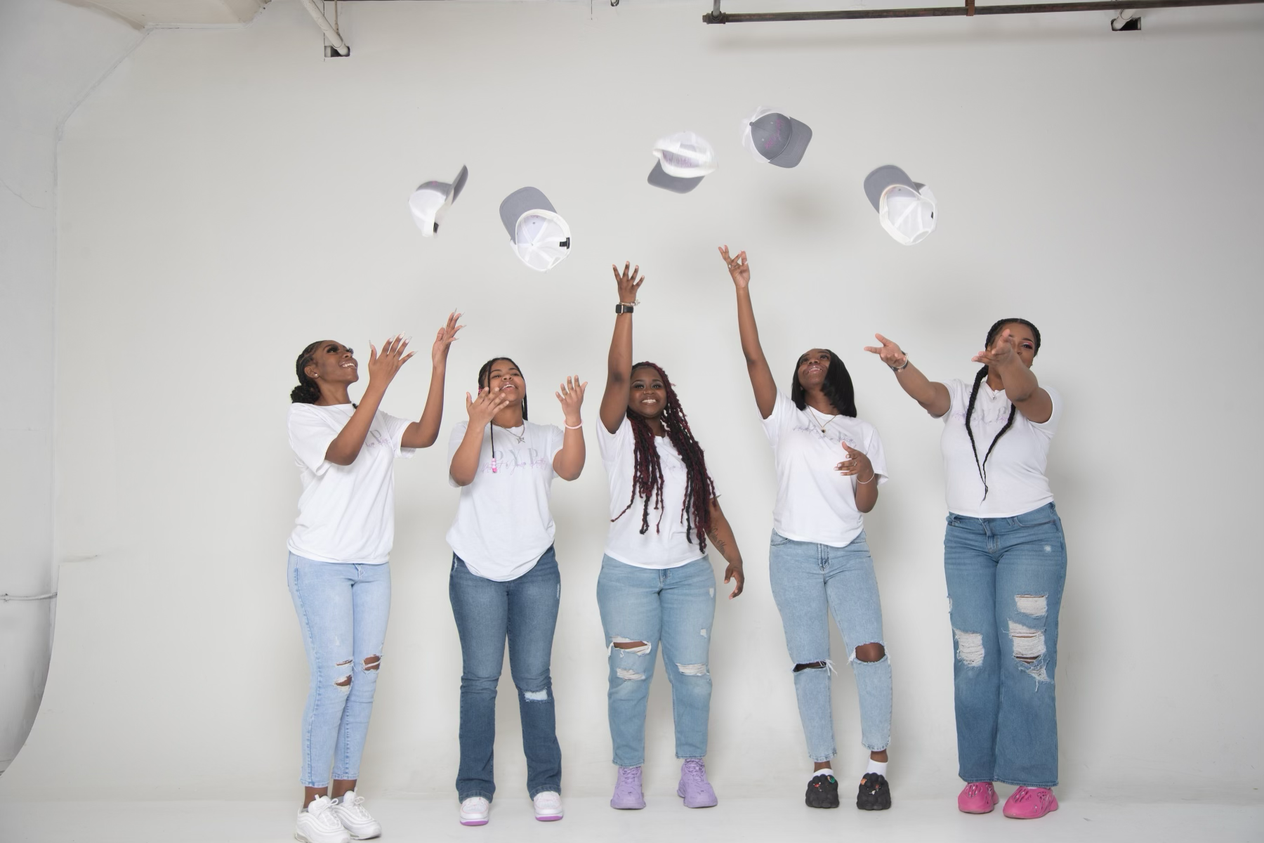 Five women wearing white t-shirts and jeans are throwing white caps into the air in a celebration against a plain white background.