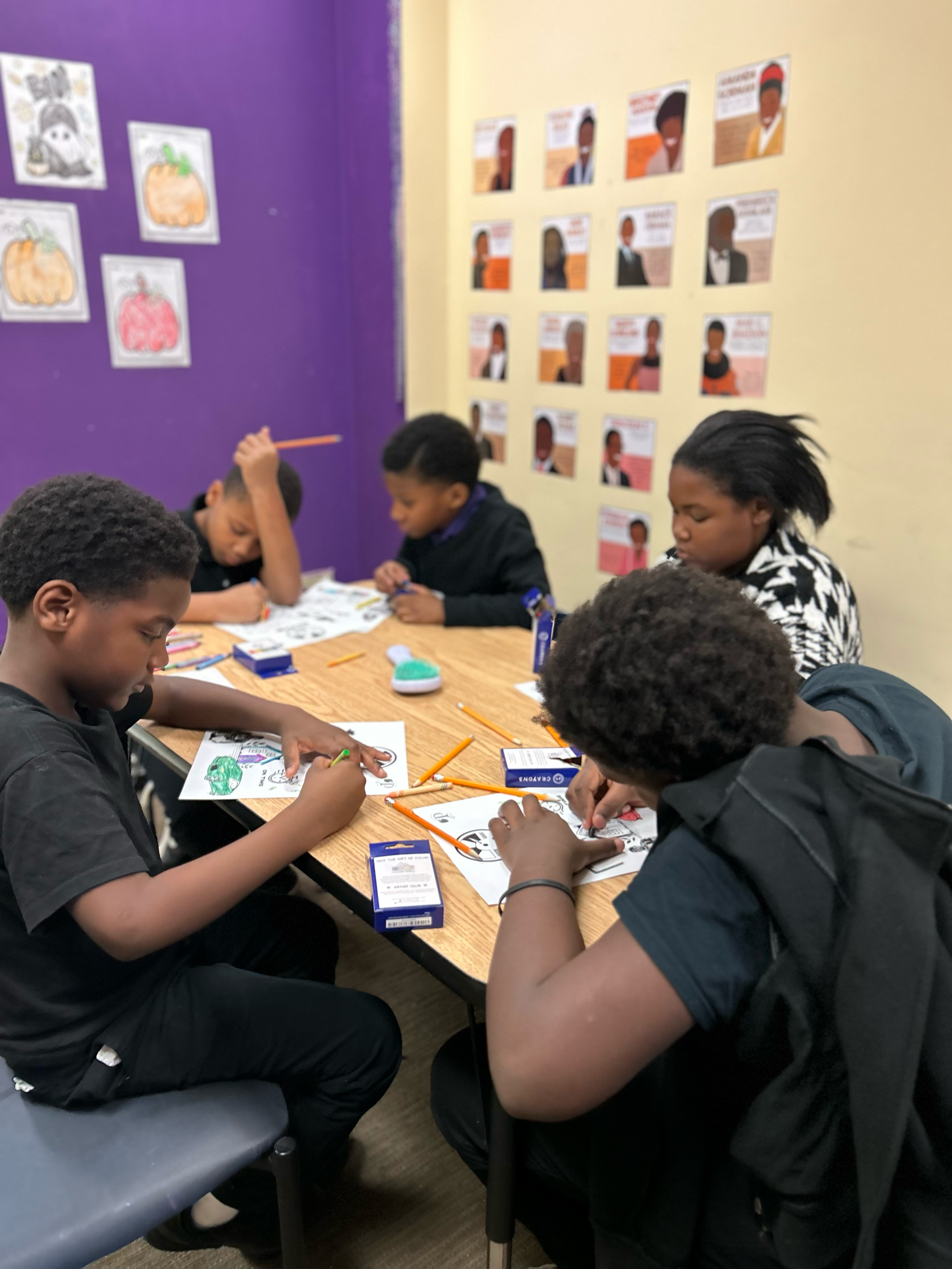 Group of five children sitting around a table coloring pictures with crayons in a classroom, with colorful posters on the walls.