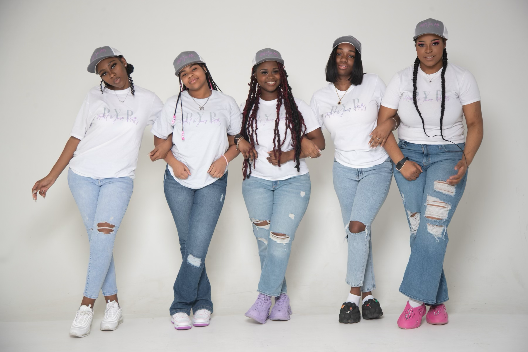 Five women standing against a white wall, wearing white T-shirts, ripped jeans, sneakers, and gray baseball caps, smiling and posing for the camera.