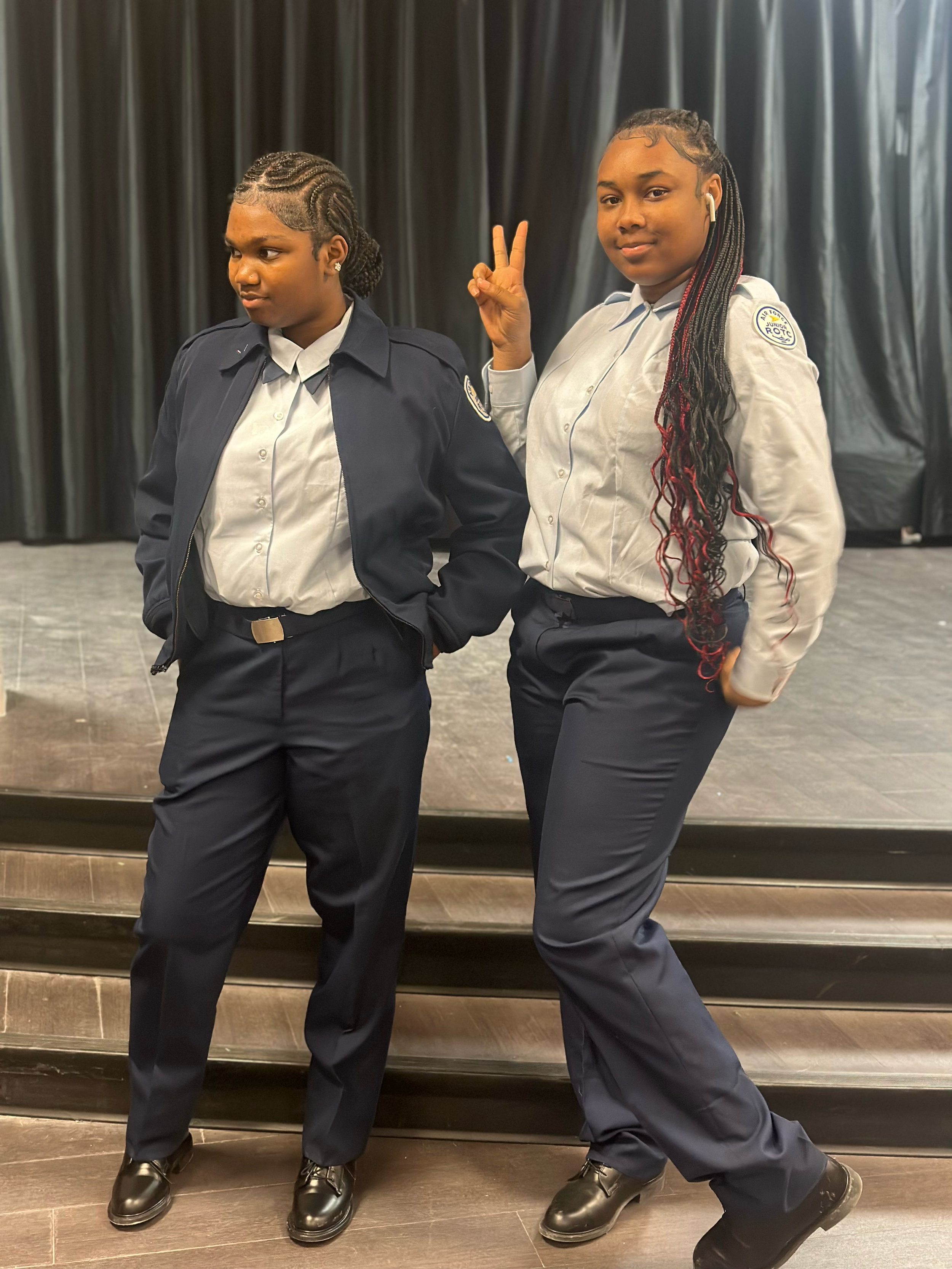 Two young women in school uniforms standing on a stage with black curtains in the background. One woman has her hands in her pockets, and the other is making a peace sign with her fingers.