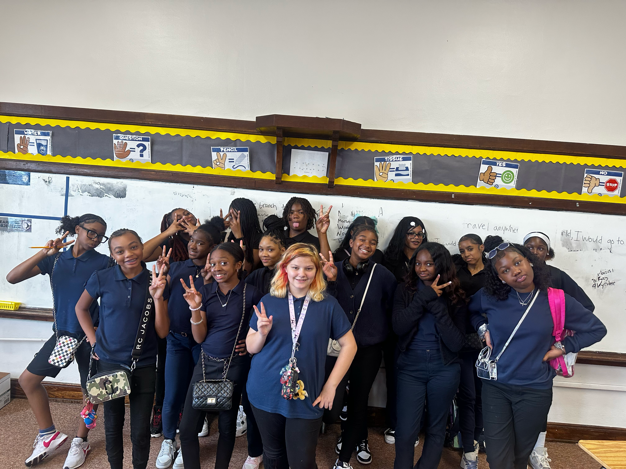 Group of young girls posing for a photo in a classroom, some making peace signs and smiling.