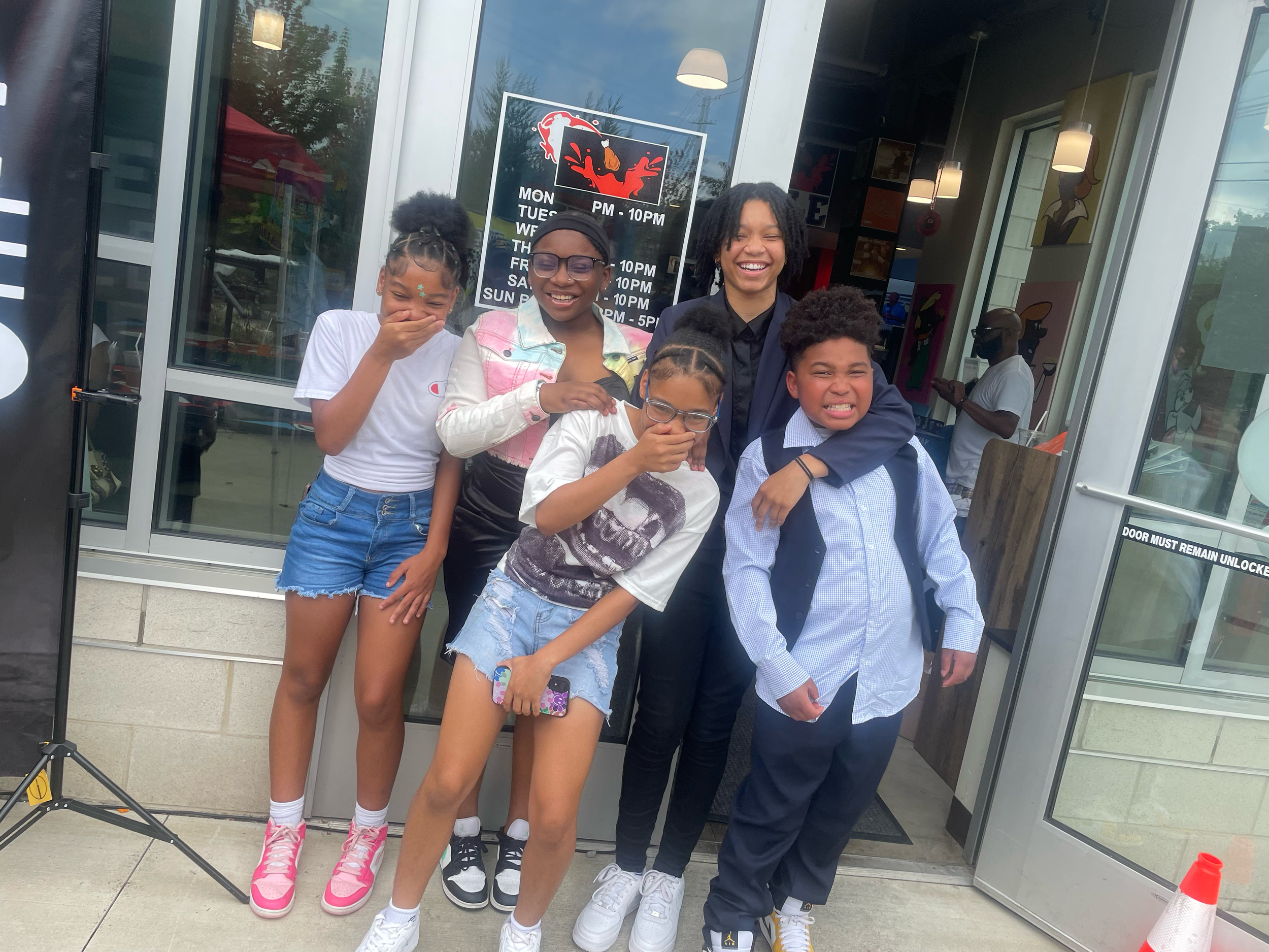 Group of five children and one adult standing in front of a restaurant entrance, laughing and making silly faces, with a person wearing a mask in the background.
