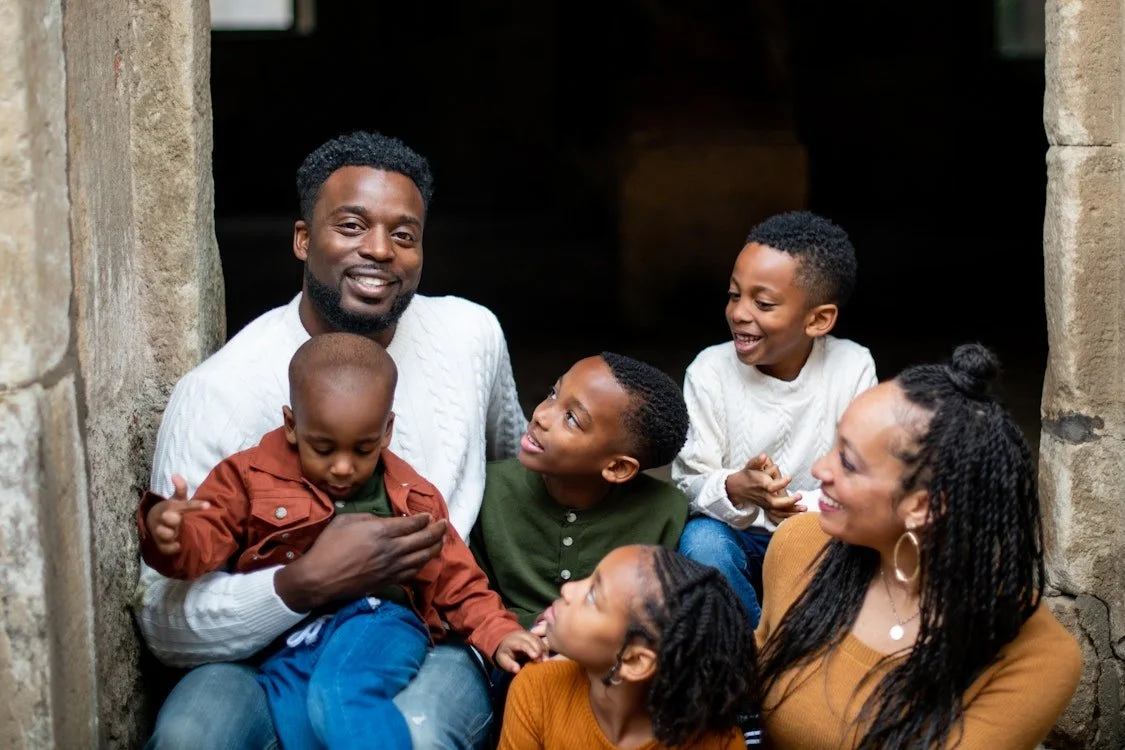 A smiling family of six, including two adults and four children, sitting together in a stone archway, enjoying each other's company.