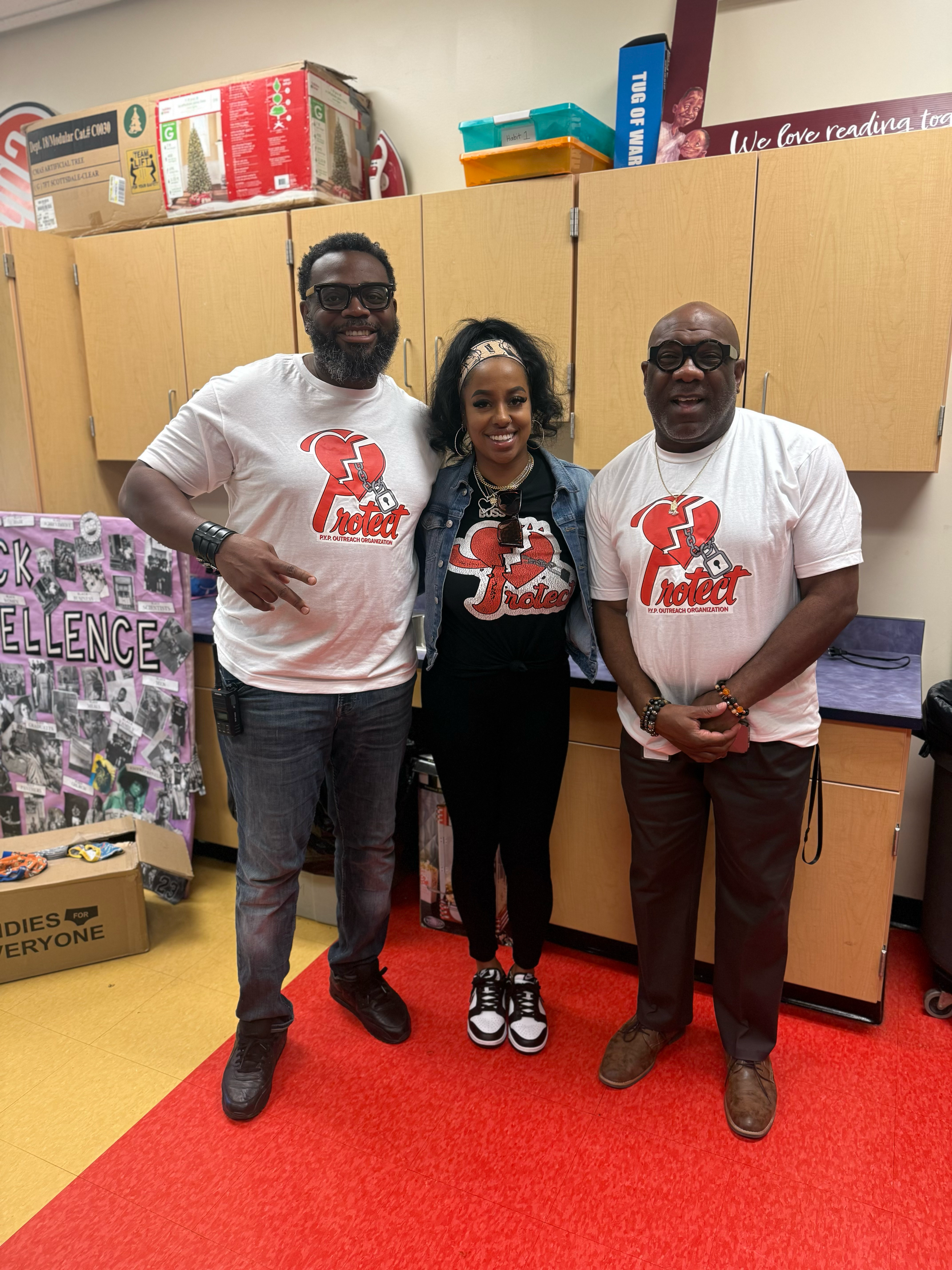 Three people standing in a room, wearing shirts with the word 'Protect' and a broken heart logo, smiling at the camera.
