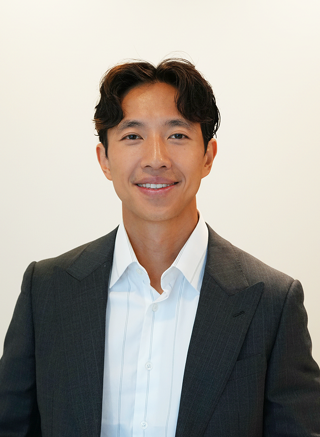 Portrait of a smiling man in a dark business suit and white dress shirt, standing against a plain white background.