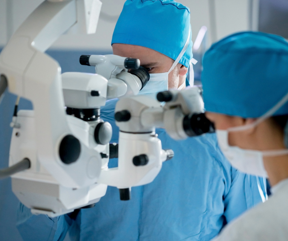 Two doctors in surgical scrubs and masks using a microscope in an operating room.