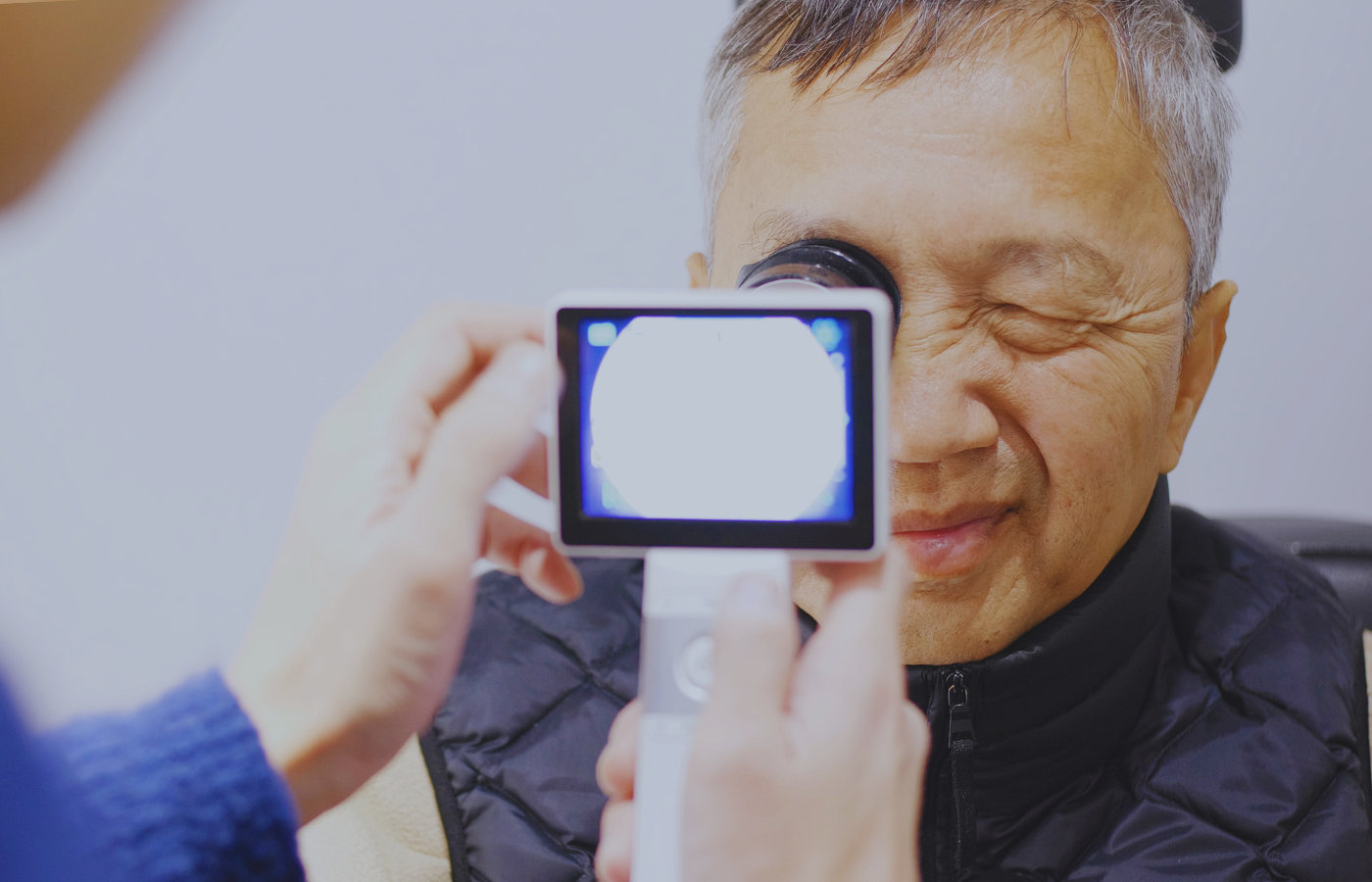 Optometrist examining an elderly man's eye using a handheld device, with the man's eye closed.