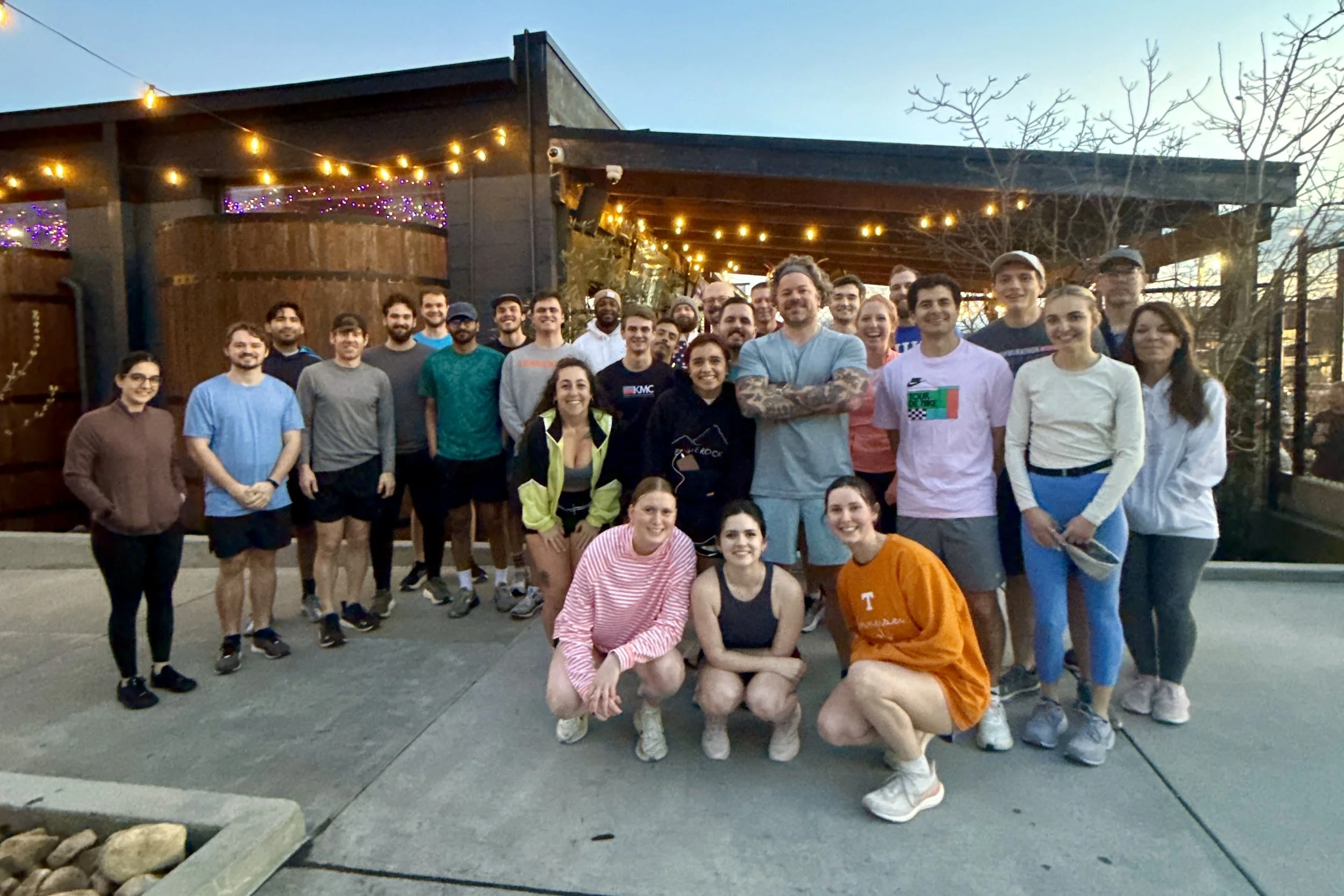 A large group of people, mostly young adults, posing outdoors in front of a building decorated with string lights and purple lights, during the evening.