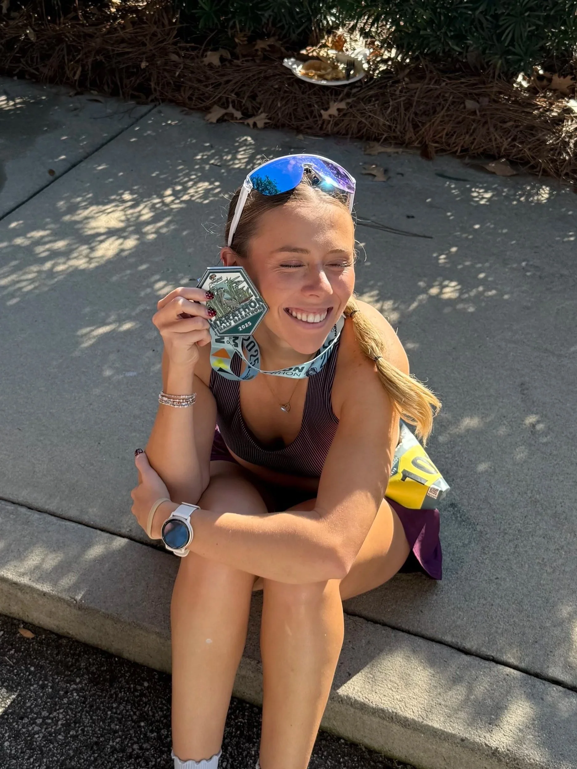 A young woman sitting on a sidewalk curb, holding a marathon medal, smiling with her eyes closed, with sunglasses on her head and a running bib around her neck, indicating she just finished a race.
