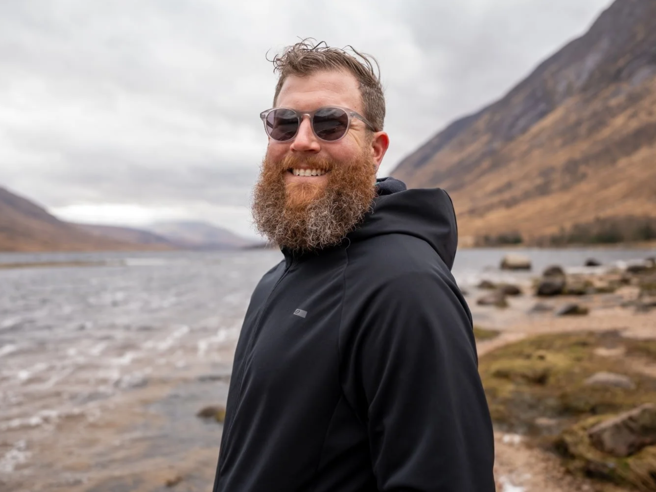 A smiling man with a beard wearing sunglasses and a black jacket standing outdoors near a river, with mountains and a cloudy sky in the background.