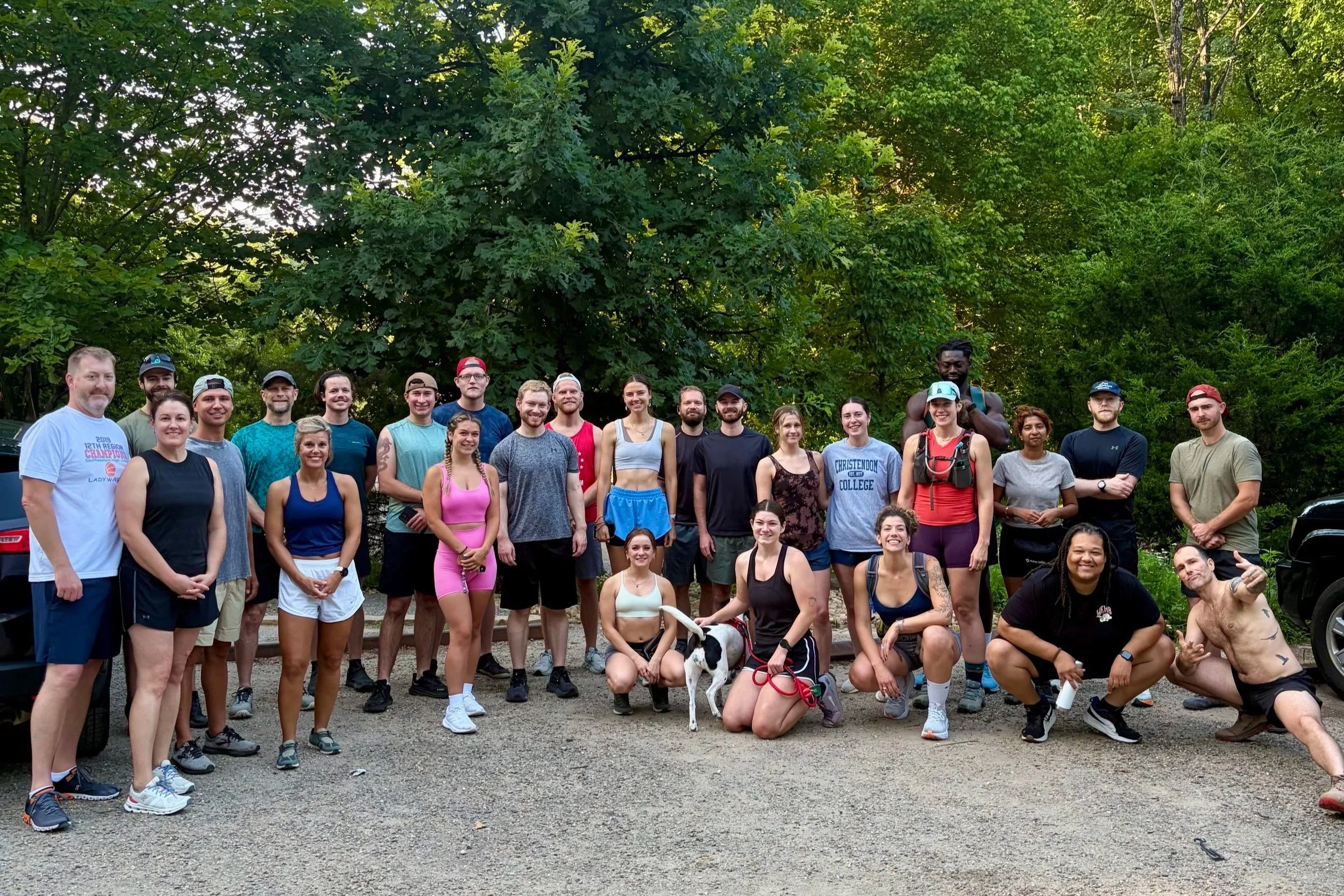 Group of diverse people standing outdoors in front of green trees, posing for a photo, with some smiling and others making playful gestures.