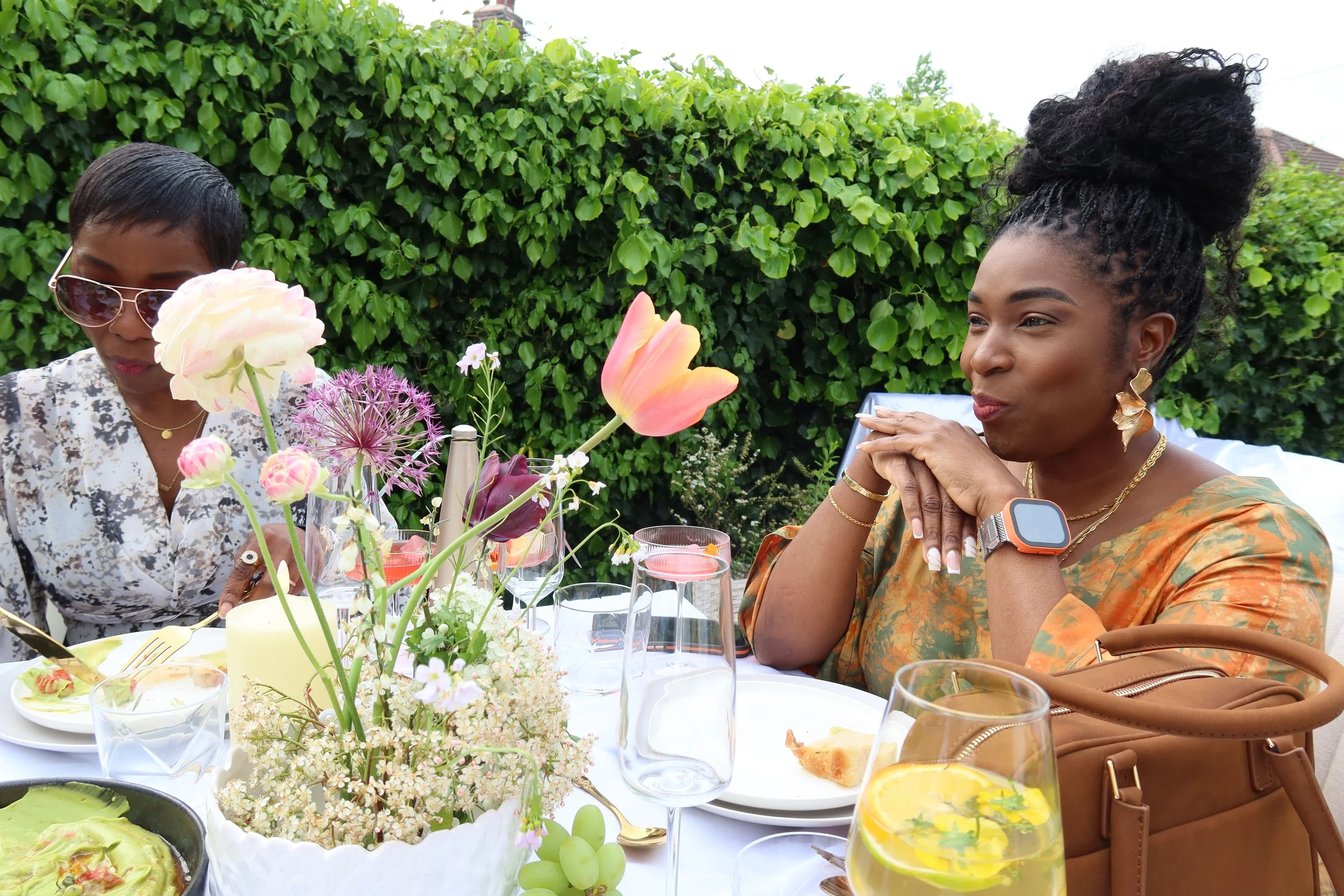 Two women sitting at an outdoor table with floral arrangements and glasses of drinks, enjoying a meal together.