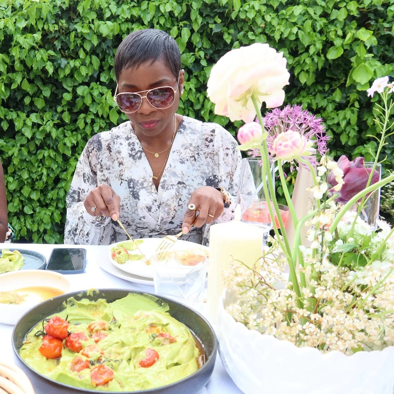 A woman wearing sunglasses and a patterned blouse at a table set outdoors with food, flowers, and greenery in the background.