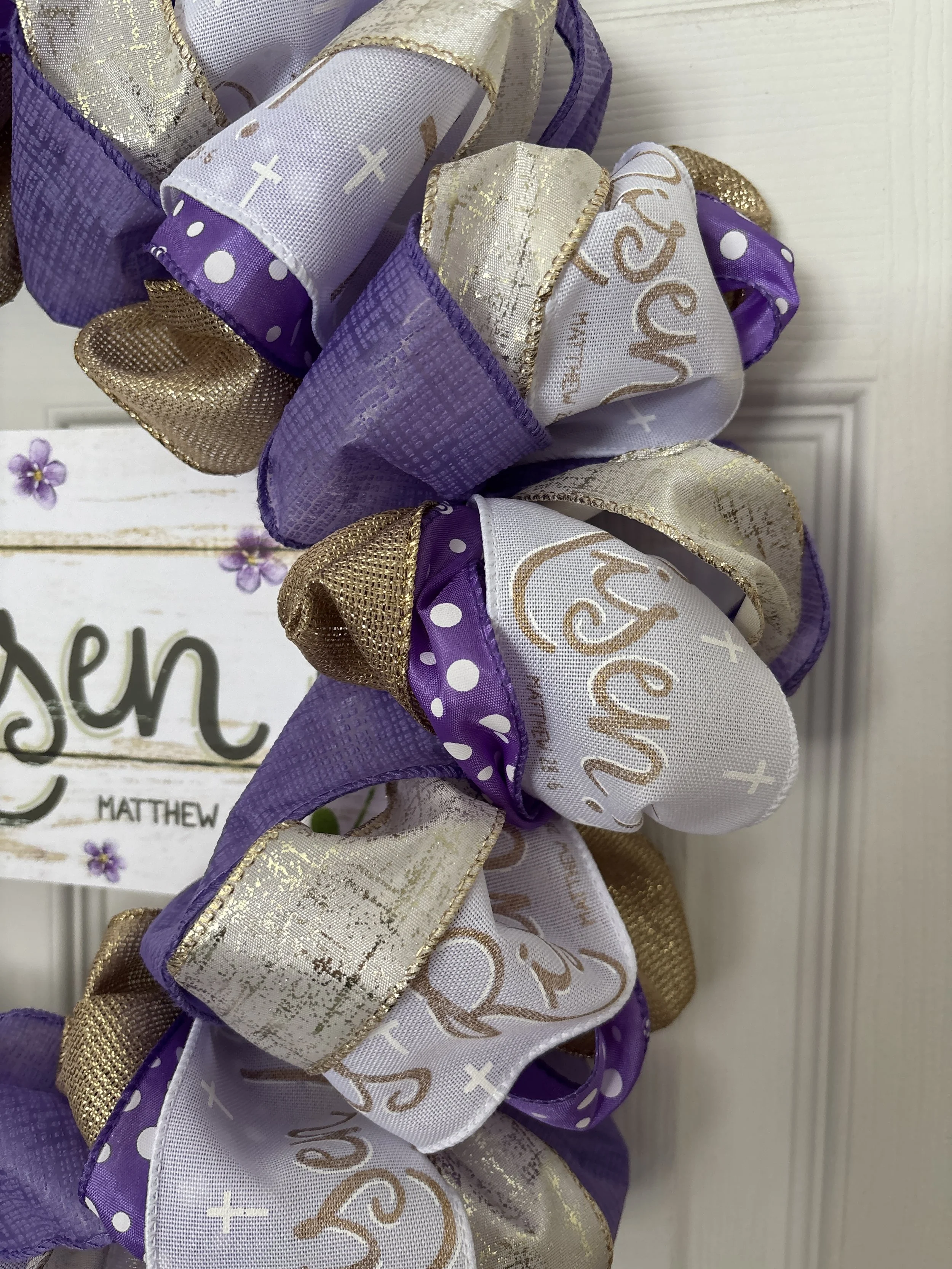 Easter wreath with white ribbon that has gold he is risen on it, purple ribbon, and gold ribbon.  White sign in the middle with gold He is Risen! letters