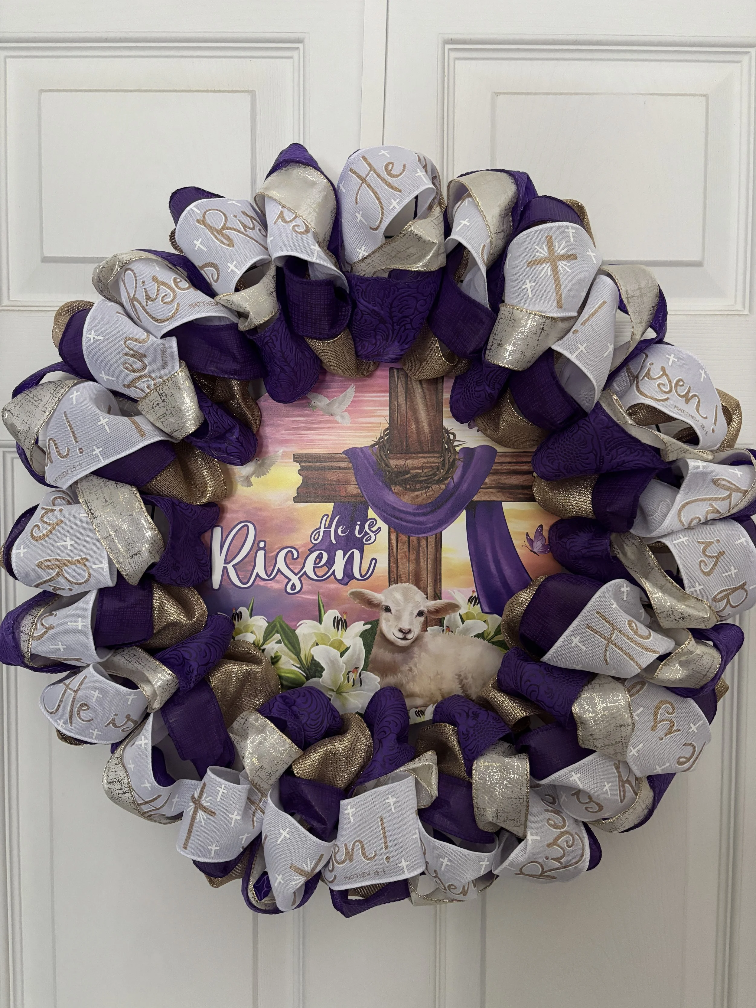 Easter wreath with purple, gold, and white ribbons, featuring the message "He is Risen" and an image of a cross with a purple cloth and a lamb.