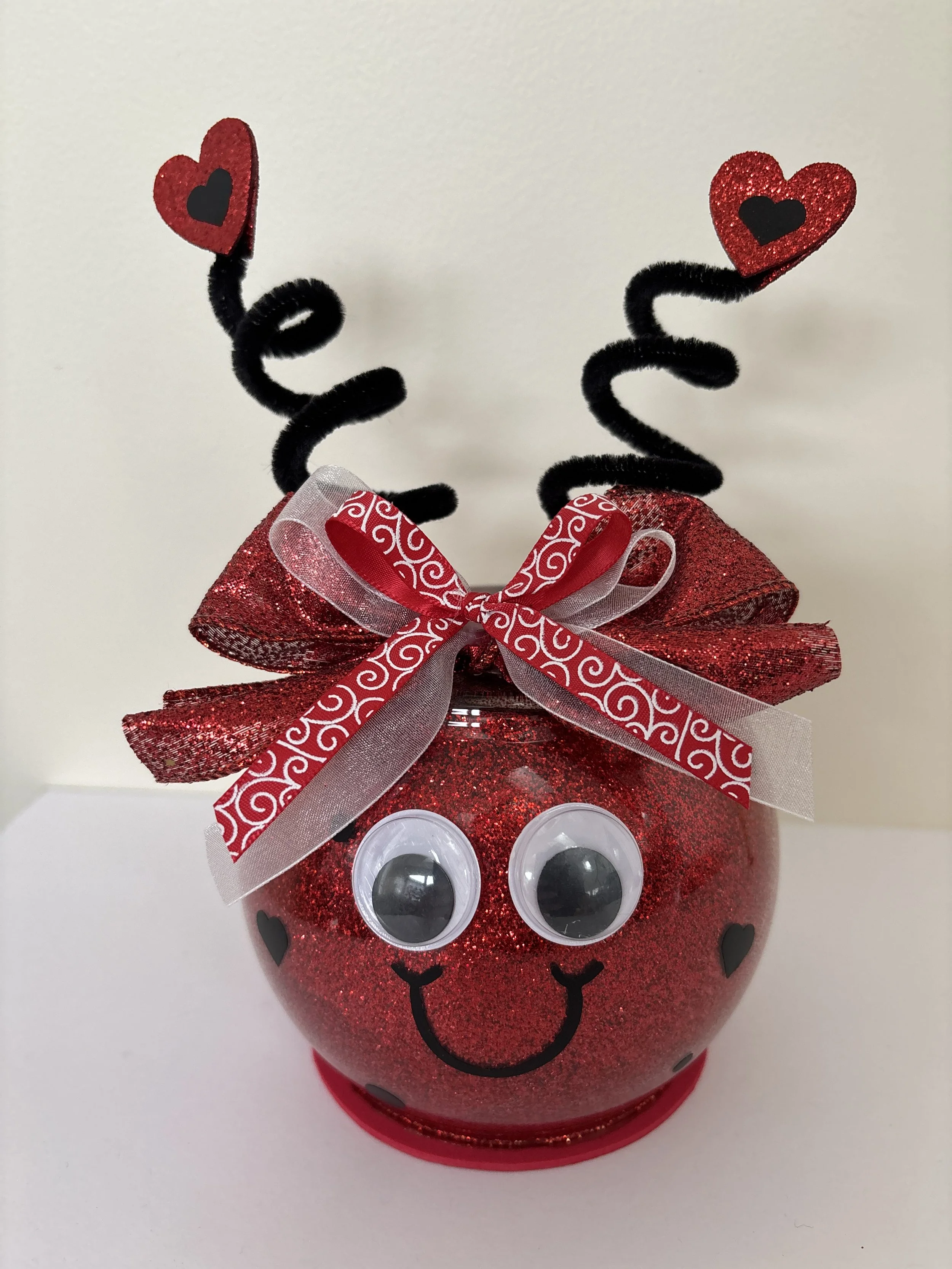 Red glass figurine with big googly eyes, red and white bow, and black antennae with hearts on them