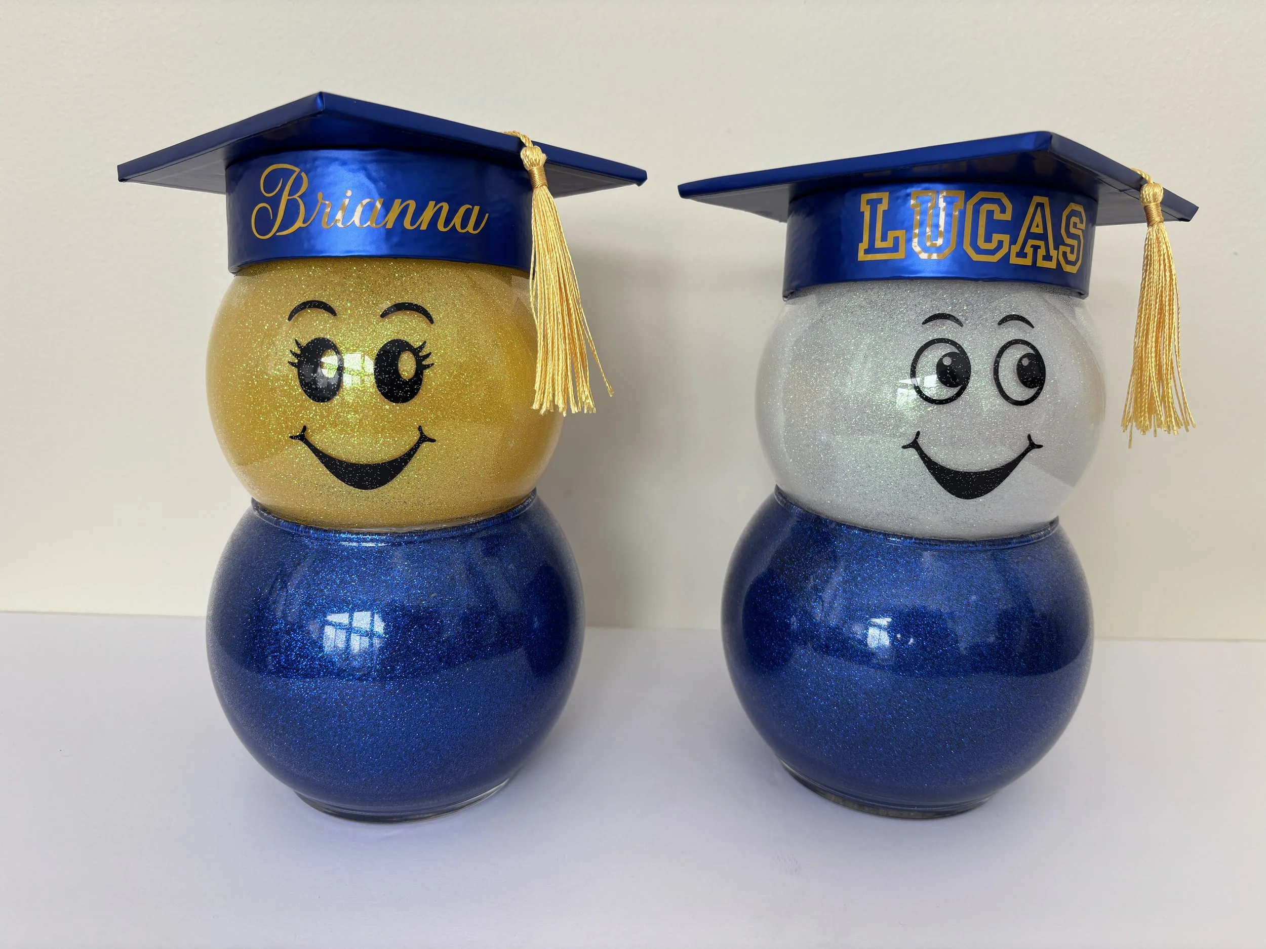 Two decorated jars with smiley face designs wearing graduation caps, one labeled 'Brianna', the other 'LUCAS'.