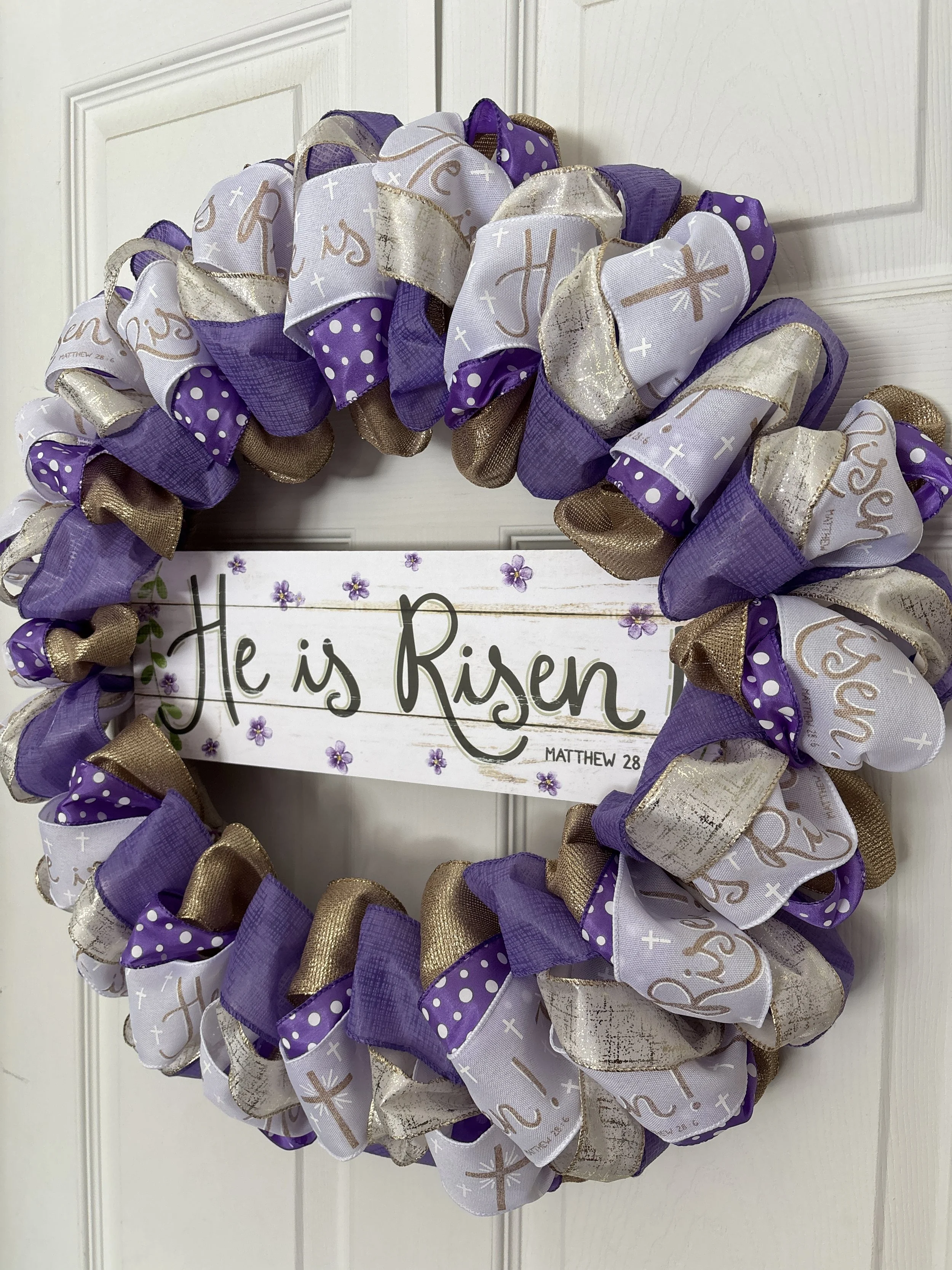 Easter wreath with white ribbon that has gold he is risen on it, purple ribbon, and gold ribbon.  White sign in the middle with gold He is Risen! letters