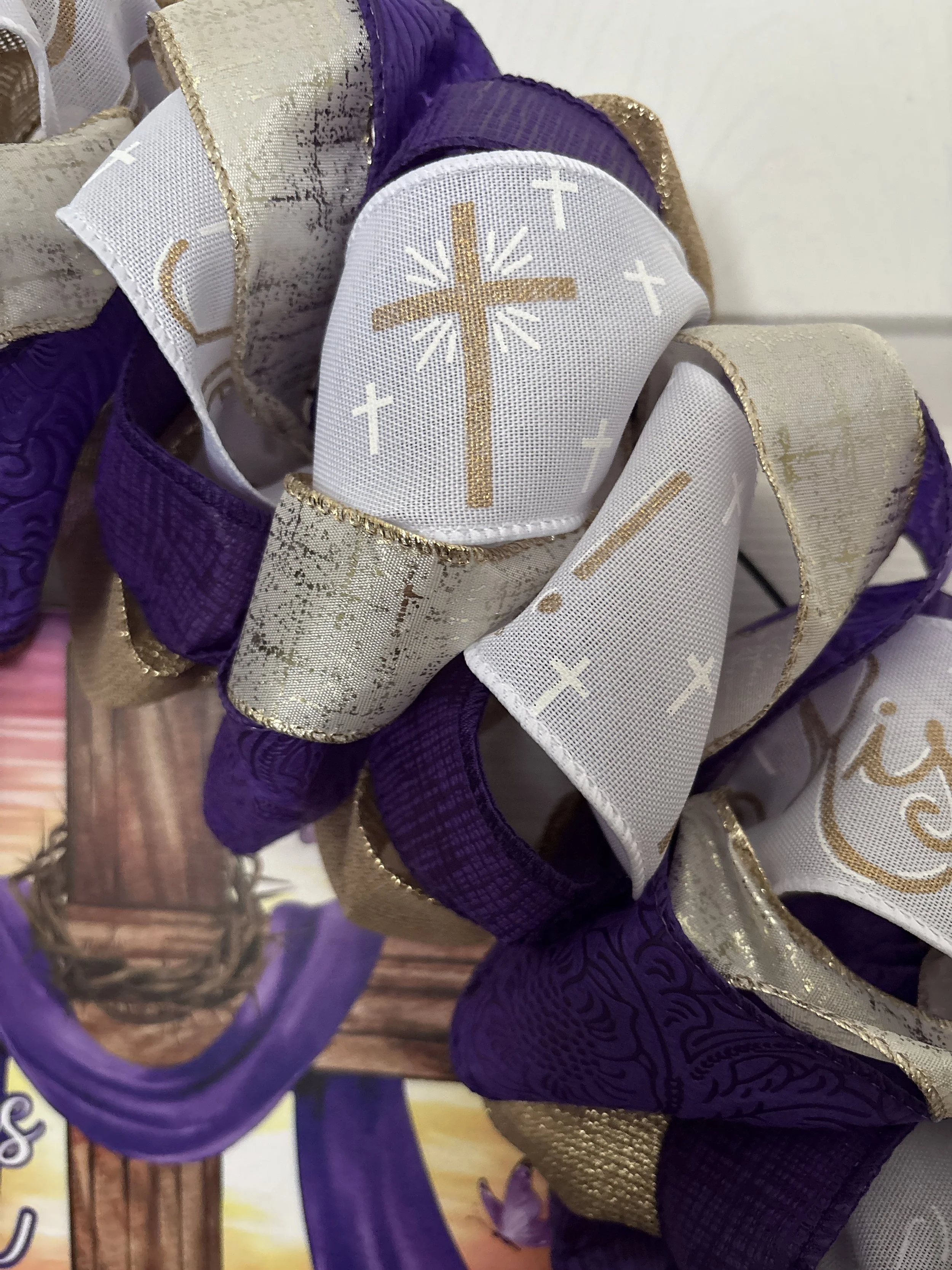 a blend of purple, gold, and white ribbons with a gold cross and “He is Risen” on the white ribbon.