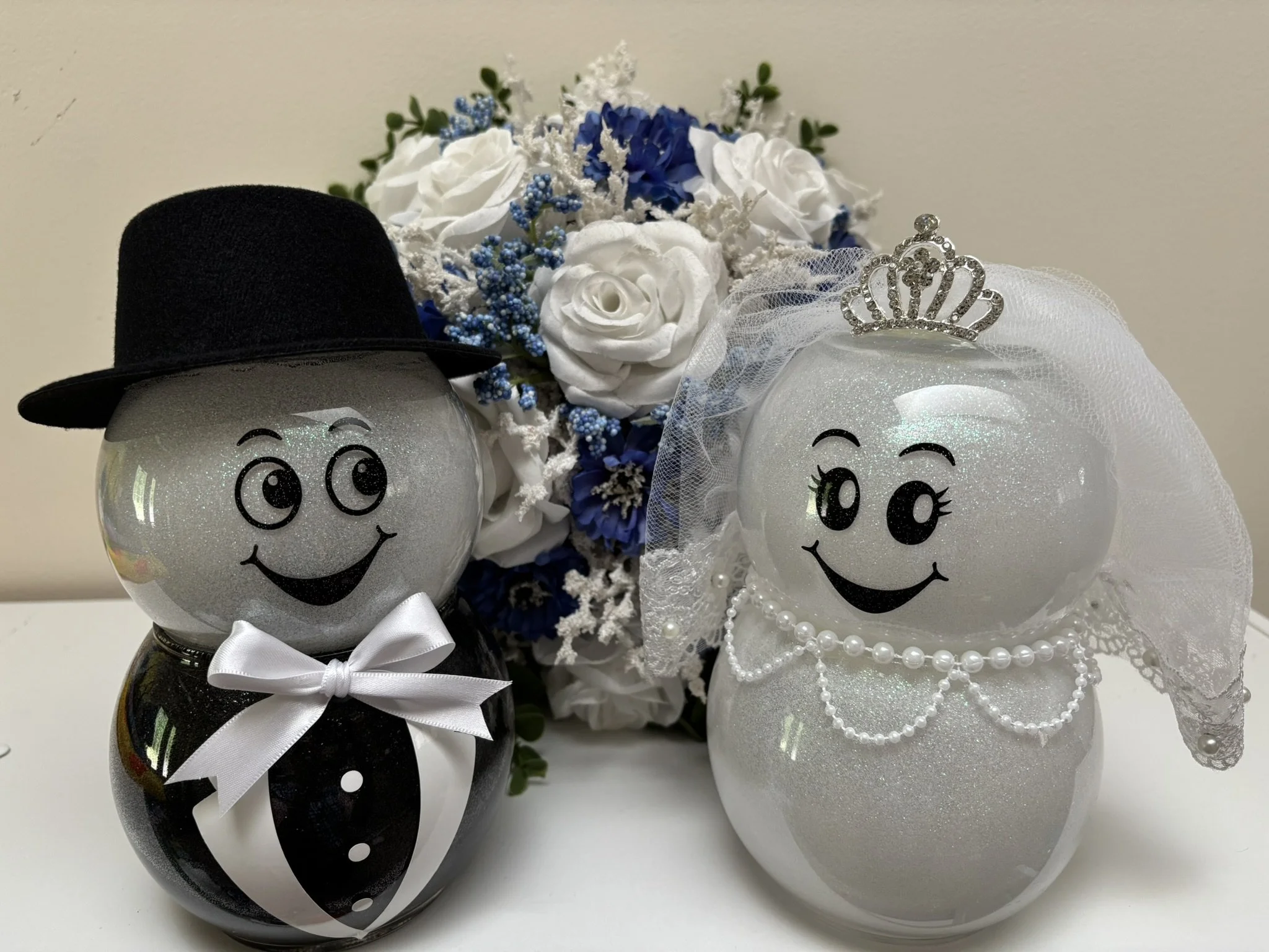 Bride and groom glass figurines.  Groom has tuxedo vest, white bow and black hat. Bride has pair of white bowls, pearl necklace, veil, and tiara.