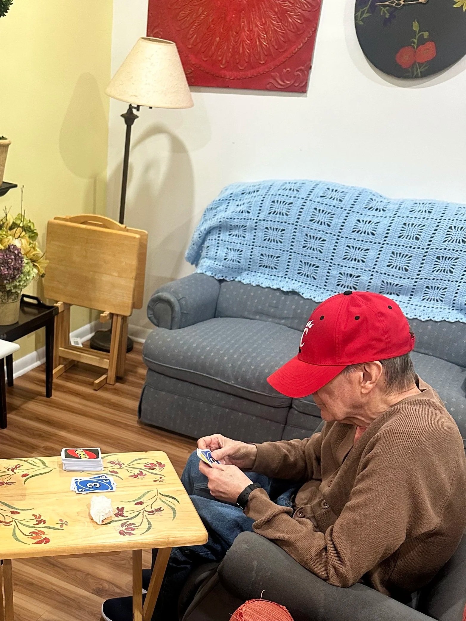 Two people sitting in a living room playing a board game, with various books and tissues on a red couch and a coffee table.