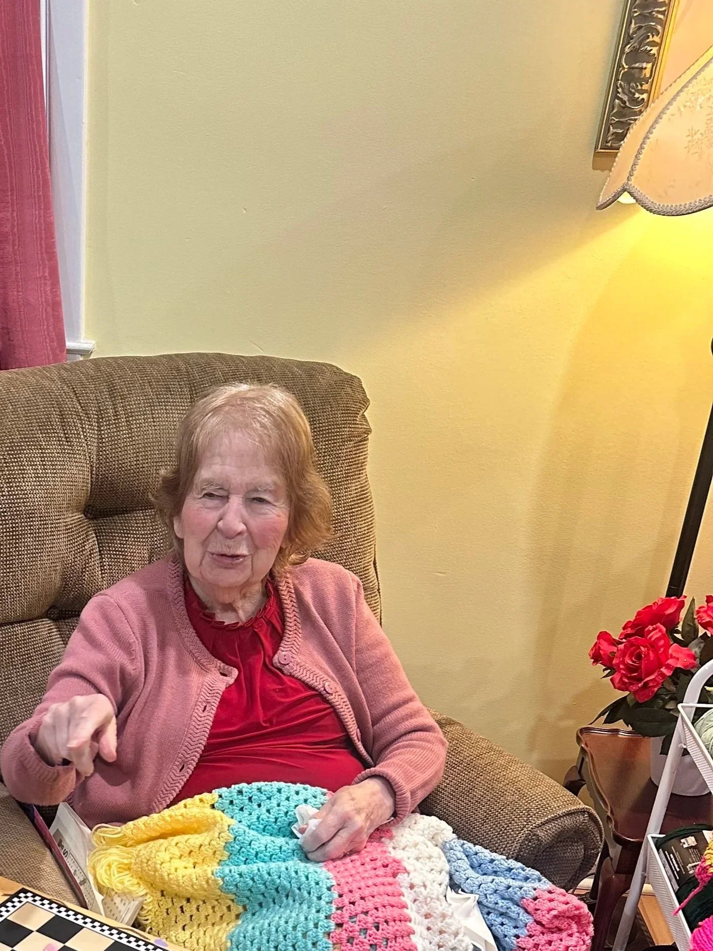An elderly woman sitting in a brown armchair, covered with a colorful crochet blanket, gesturing with her right hand while talking to a young person playing checkers. The young person, wearing a blue shirt and sneakers, sits at a small table with a c