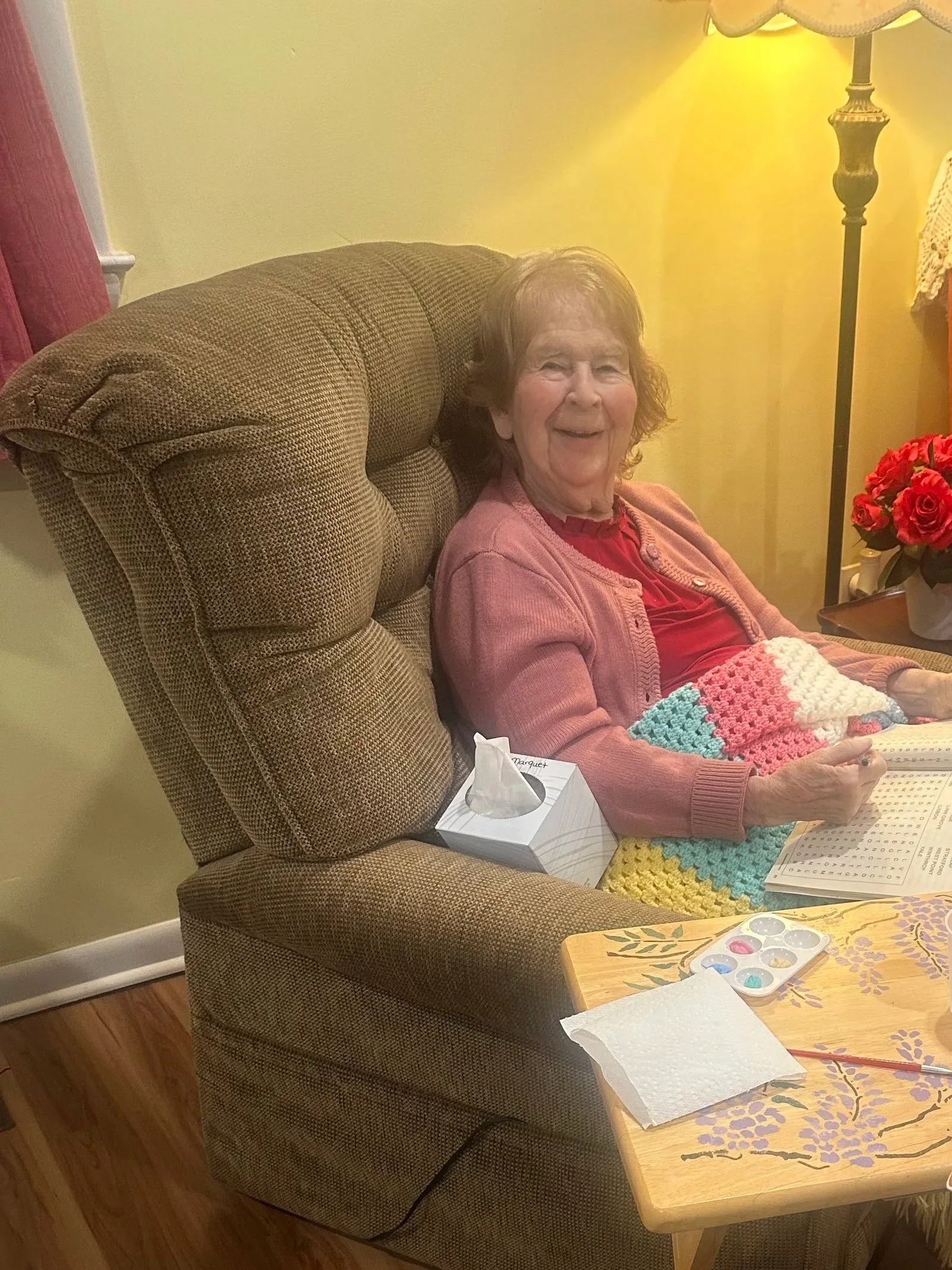 An elderly woman with short curly hair, smiling and sitting in a cushioned armchair, covered with a multi-colored crocheted blanket. There is a man in a light blue shirt and black pants sitting nearby, holding a sunny-side-up egg on a plate. A small 