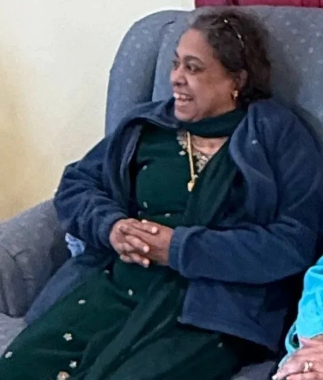 Two women sitting on a gray couch in a living room, smiling and engaging in conversation, with decorative curtains behind them and wooden flooring.