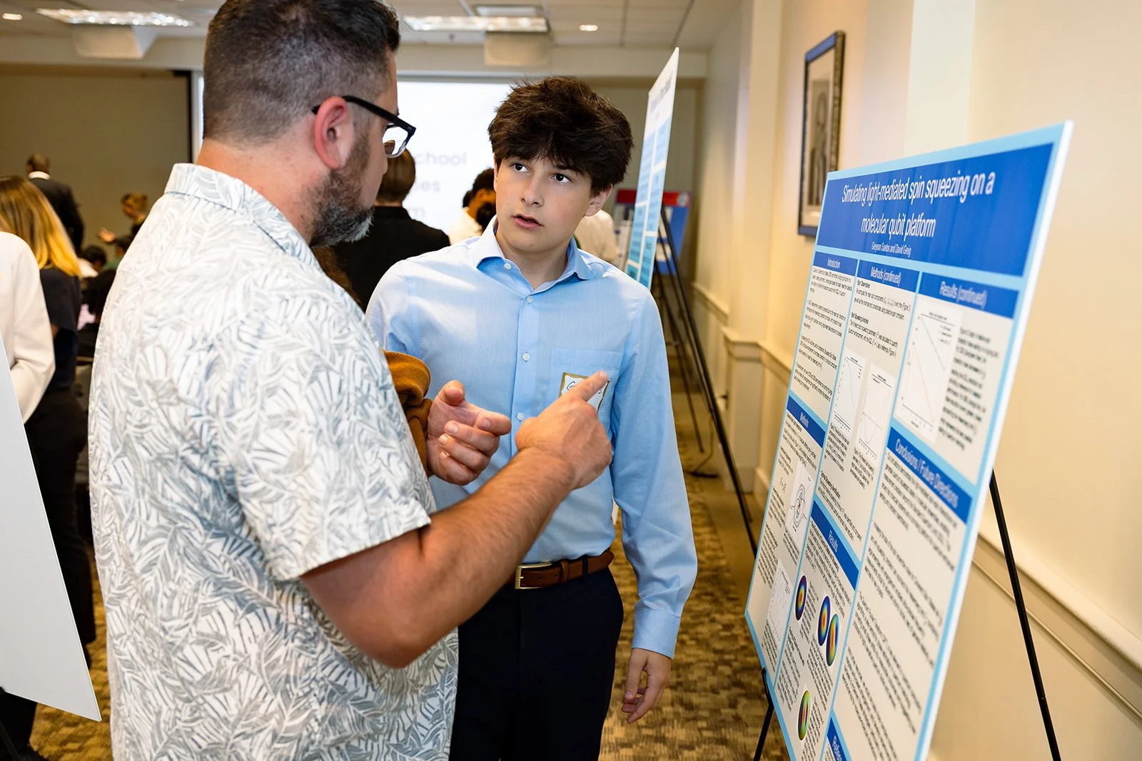 Two men, one older with glasses and a patterned shirt, and a younger man in a blue shirt, are engaged in conversation at a scientific conference. The older man is pointing at a research poster titled "Synthetically light-modulated spin squeezing on a molecular qubit platform" displayed on an easel.