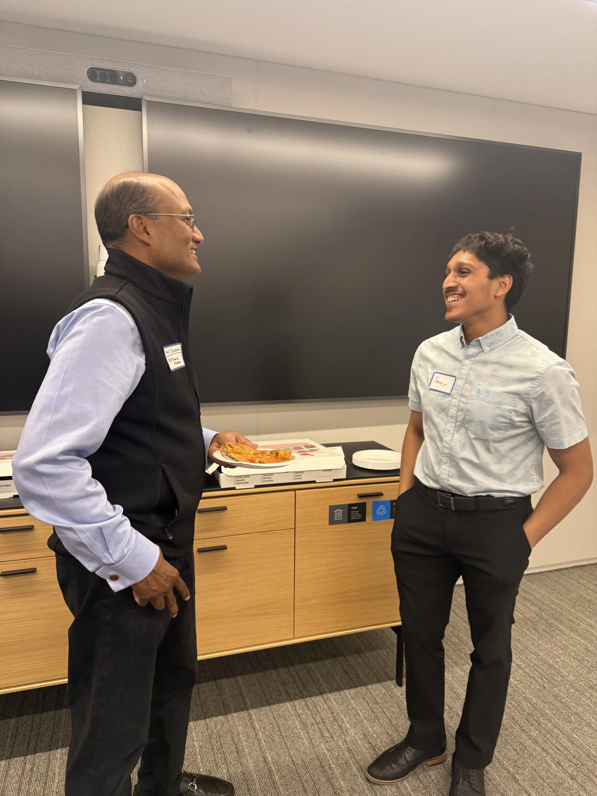Two men are having a conversation in an office break room, with one holding a slice of pizza. They are smiling and dressed in business casual attire.