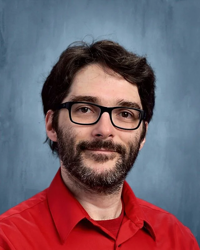 A man with dark hair, glasses, and a beard, wearing a red shirt against a blue background.