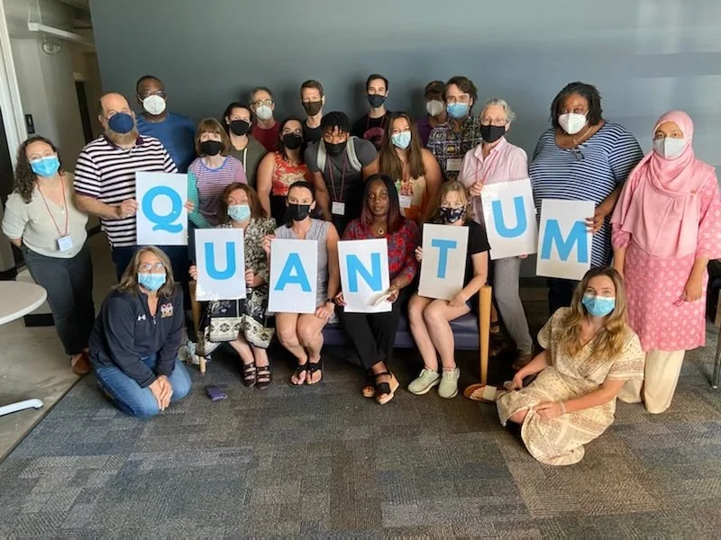 Group of diverse people holding large letters spelling 'QUANTUM' during an indoor event, all wearing face masks.