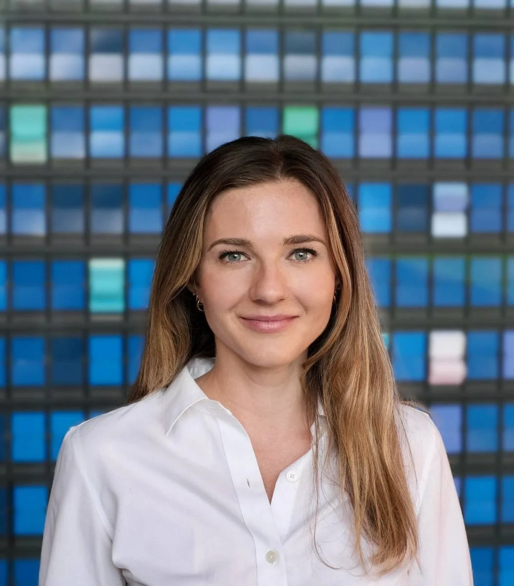 A woman with light brown hair, wearing a white button-up shirt, standing in front of a building with a multi-colored glass facade.