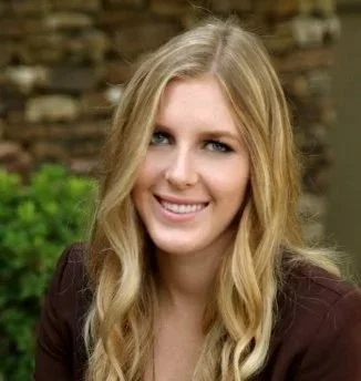 A young woman with long blonde hair, smiling outdoors in front of a brick wall and greenery.