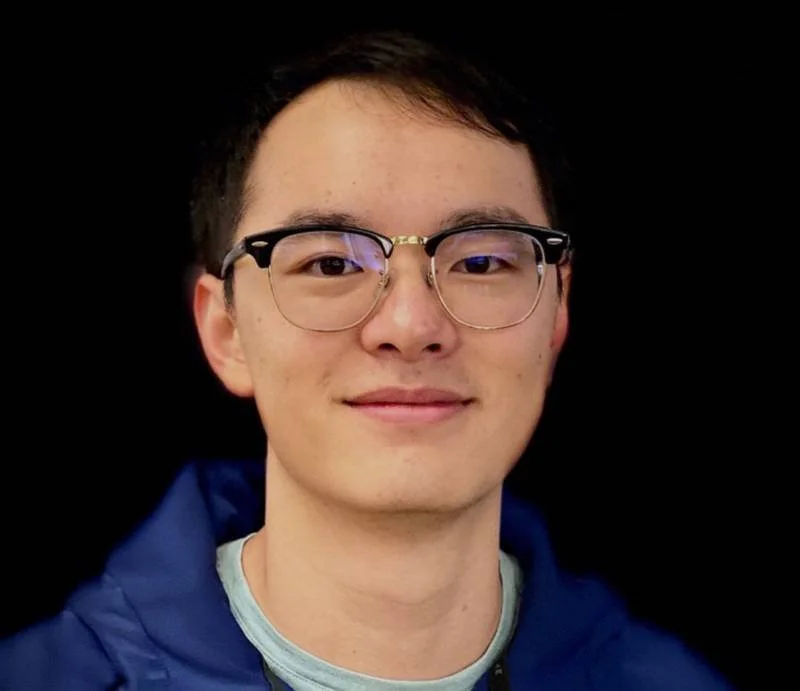 Young man with short dark hair, glasses, wearing a dark blue hoodie, smiling in front of a black background.