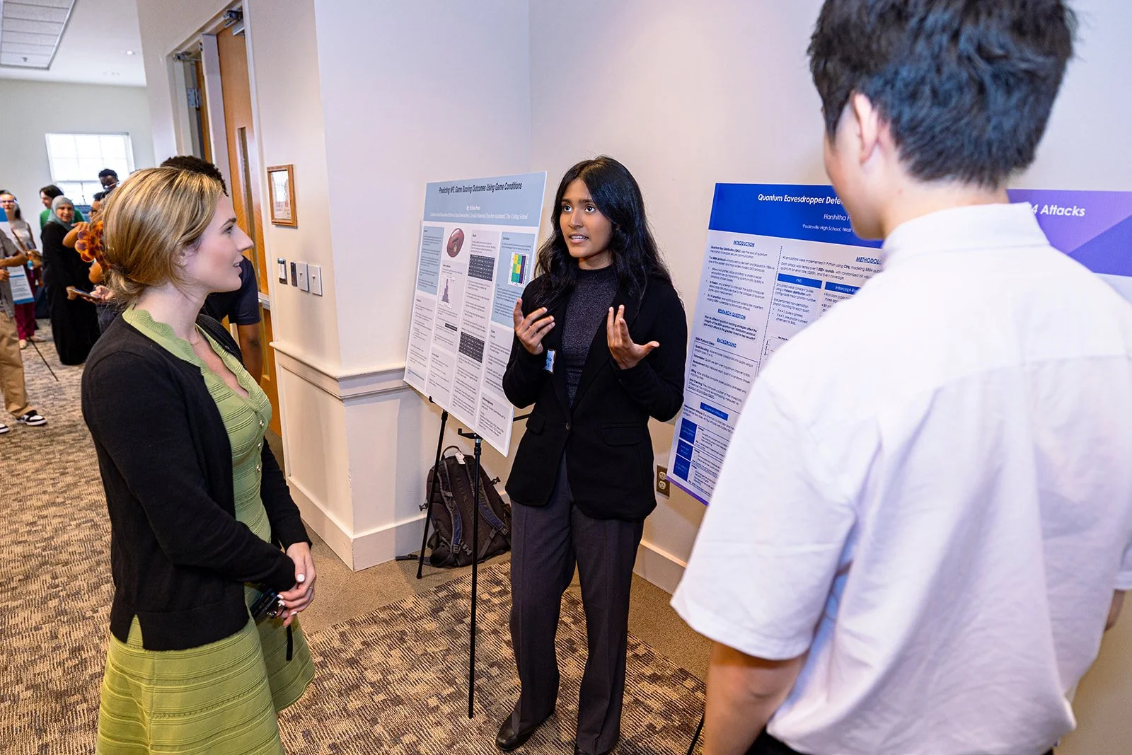 A woman in business attire is giving a presentation at a research poster session while two people listen. The setting is indoors, with other attendees and posters visible in the background.